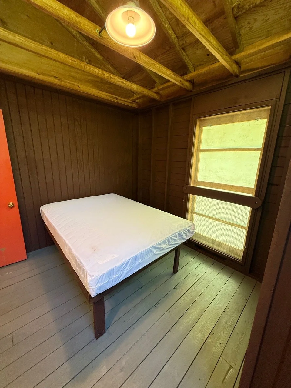 Empty bedroom with wood-paneled walls, single bed with mattress, wooden floor, window with shades, and exposed wooden ceiling beams.