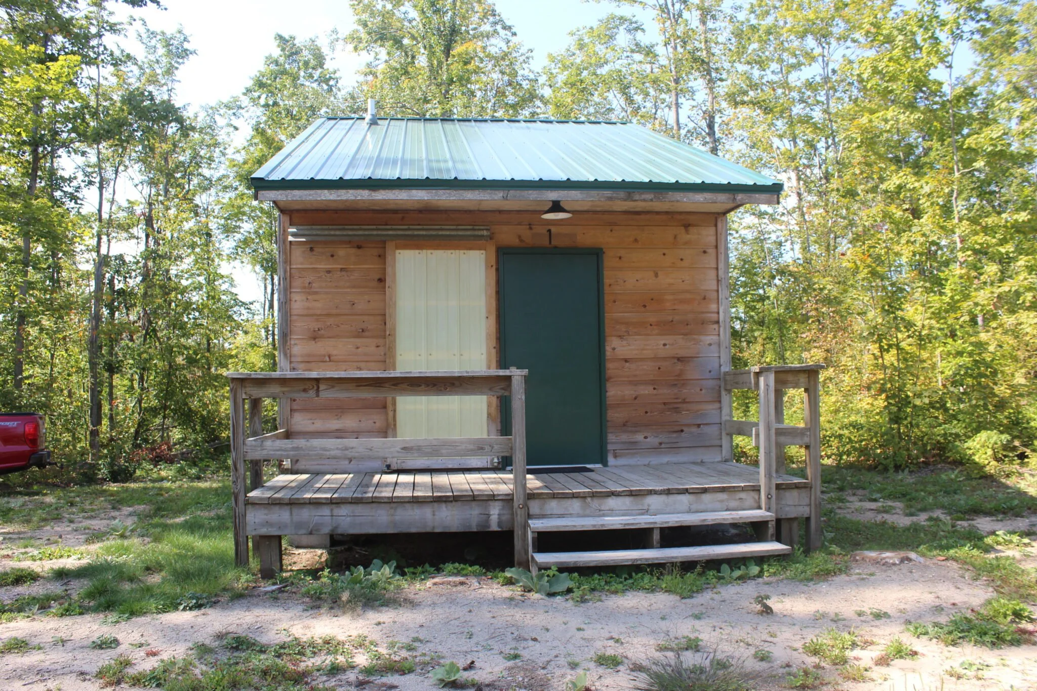 Small wooden cabin with green metal roof, green door, and beige window, surrounded by trees with green leaves, and a small deck with railings in a forested area.