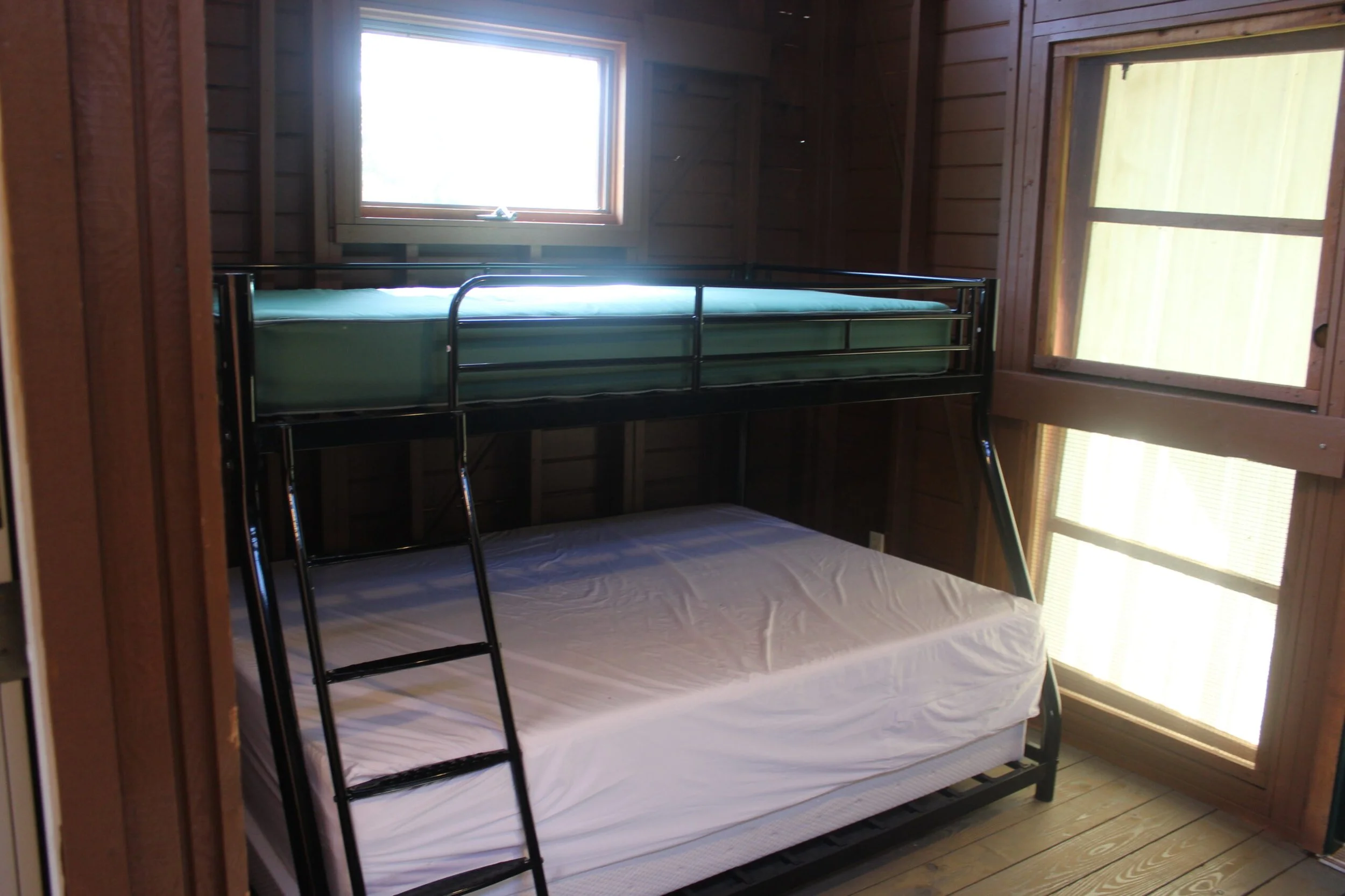 A bunk bed setup inside a wooden room with large windows. The top bunk has a metal frame with a mattress, while the bottom bunk has a mattress but no bedding.
