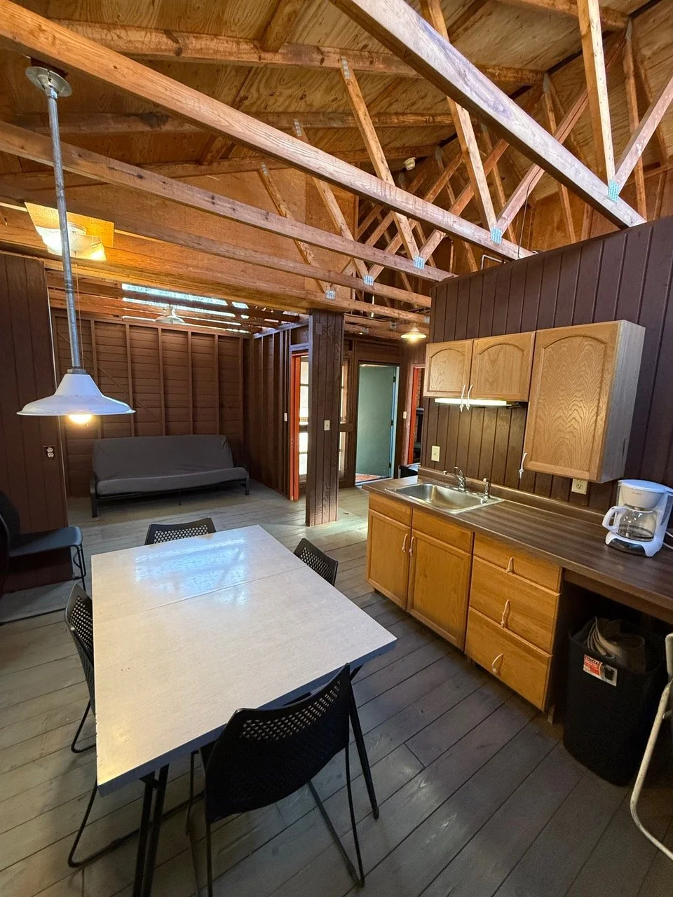 Interior of a rustic-style cabin kitchen with wooden walls, ceiling beams, a dining table with black chairs, a kitchenette with cabinets, a sink, a coffee maker, and a futon in the background.