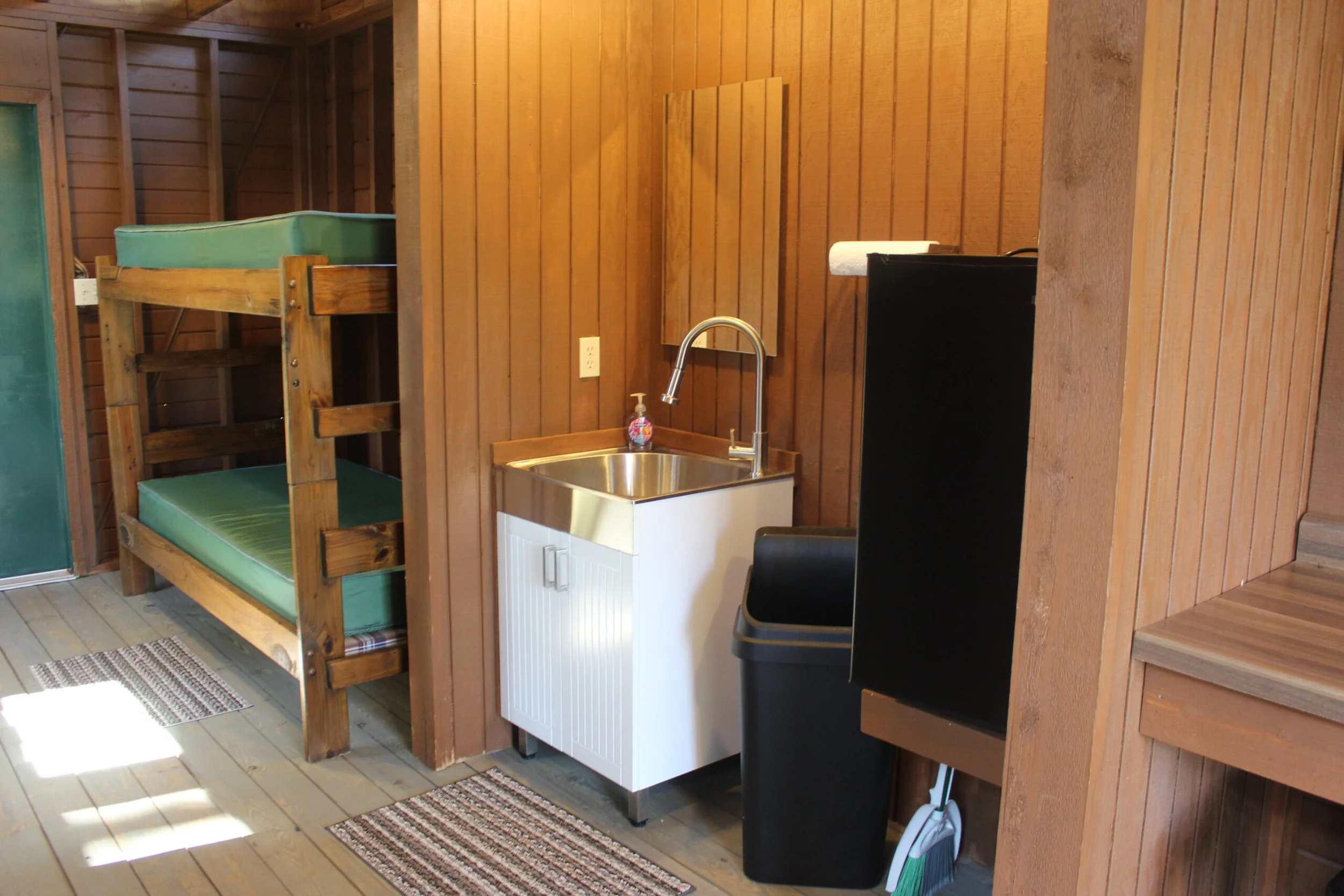 Interior of a wooden cabin or tiny house with bunk beds, a small sink, a mini fridge, and part of a staircase leading up to another level.
