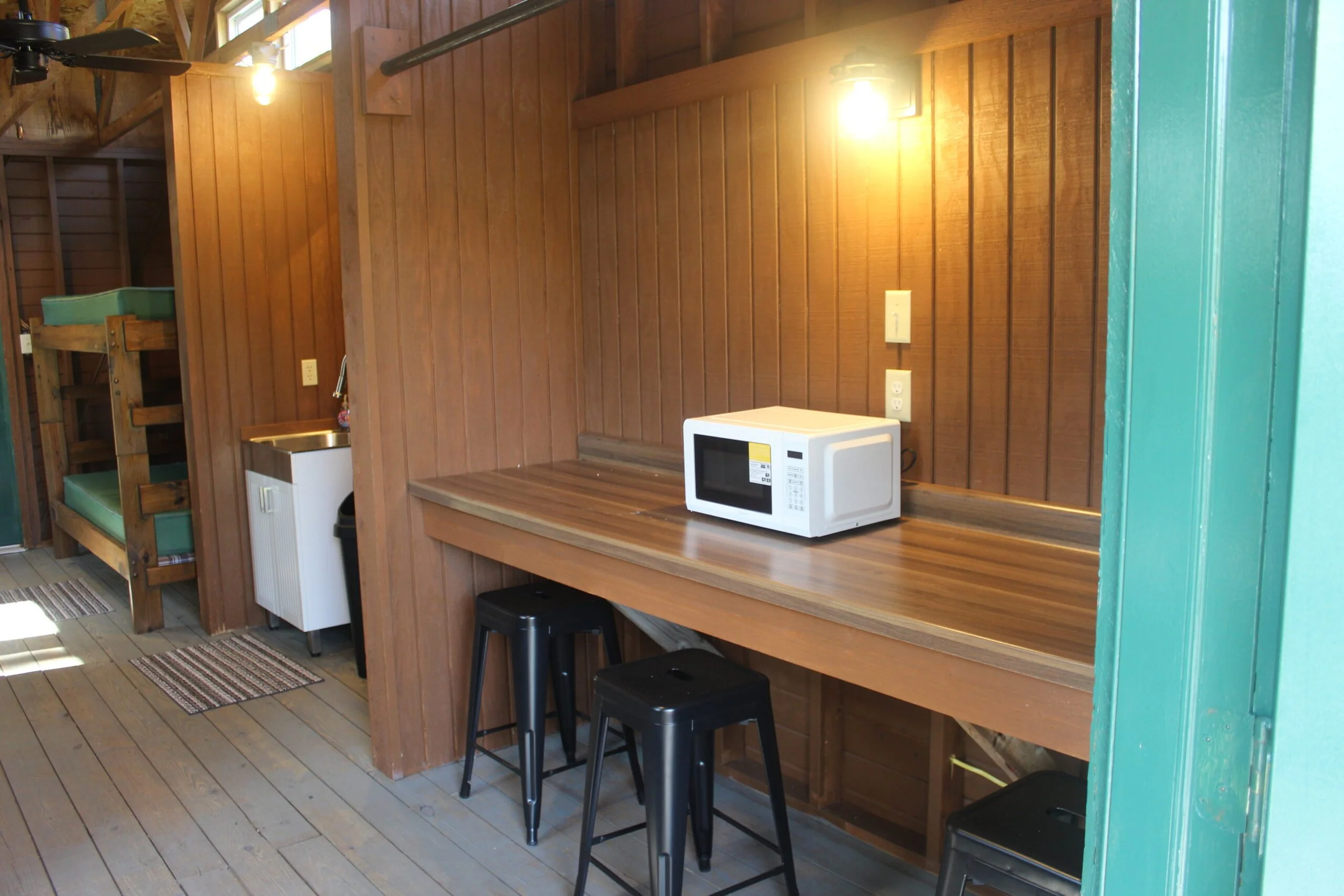 Interior of a wooden cabin with a microwave on a wooden counter, black stools, a small sink, and wooden walls and ceiling. There is a ceiling fan and windows letting in natural light.