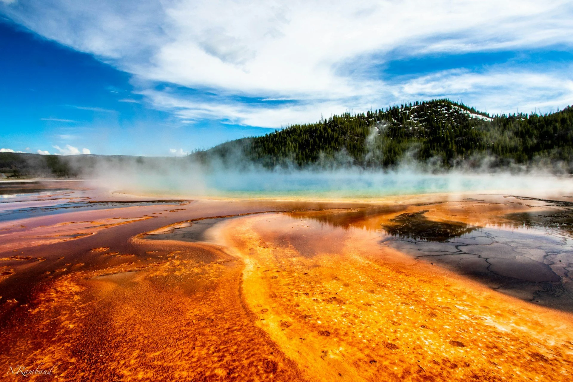 Colorful geothermal hot spring with steaming water, surrounded by orange, yellow, and black mineral deposits, with a forested hill and cloudy sky in the background.