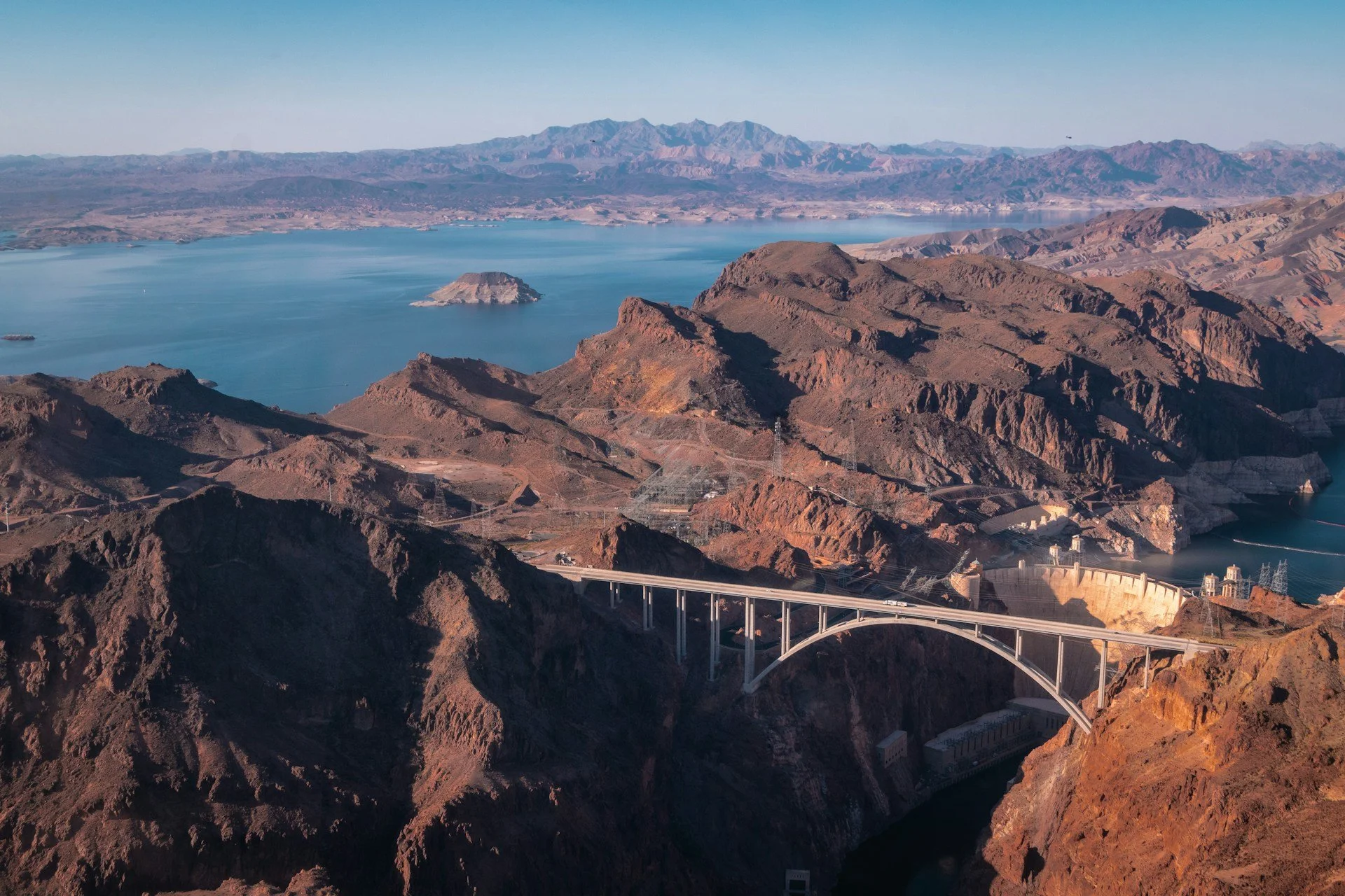Aerial view of the Hoover Dam with surrounding rugged cliffs and Lake Mead in the background. Mountain ranges extend into the distance under clear blue skies.