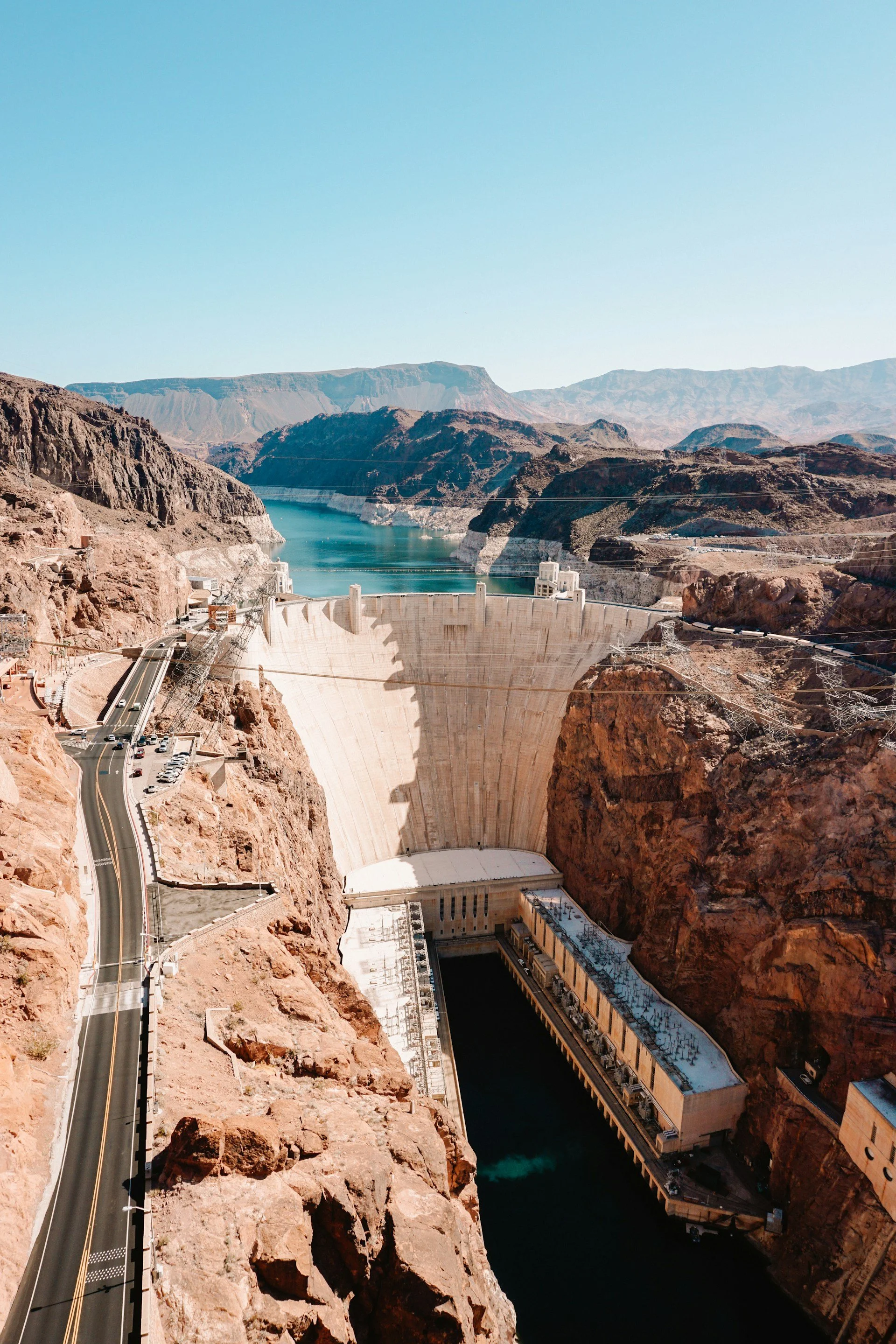 The image shows the Hoover Dam situated in the rocky landscape of the Black Canyon with a highway and parked cars on the left side. The dam spans the Colorado River, with water behind it and mountains in the background under a clear blue sky.