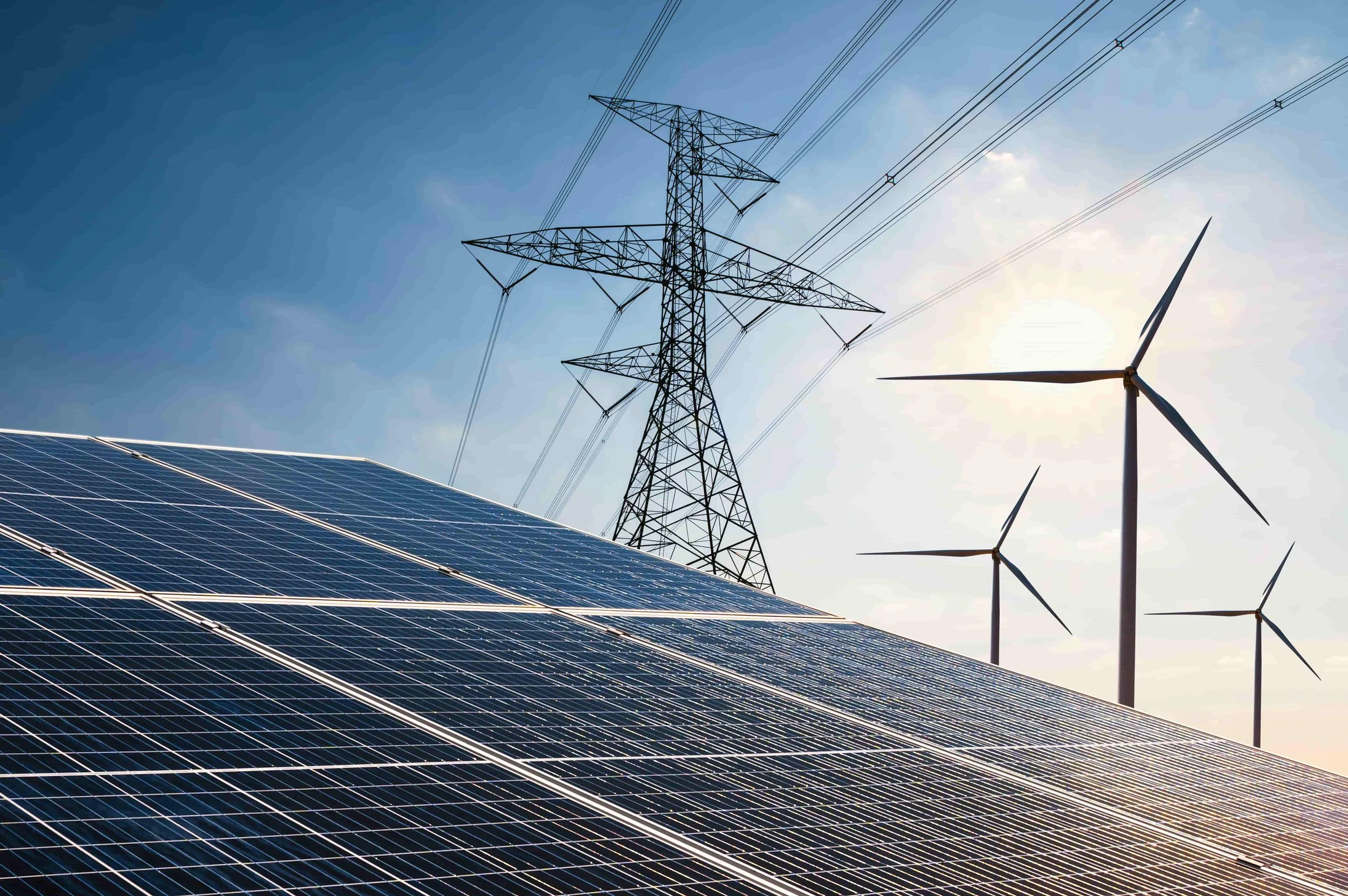Solar panels, a transmission tower with power lines, and a wind turbine against a blue sky with sunlight.