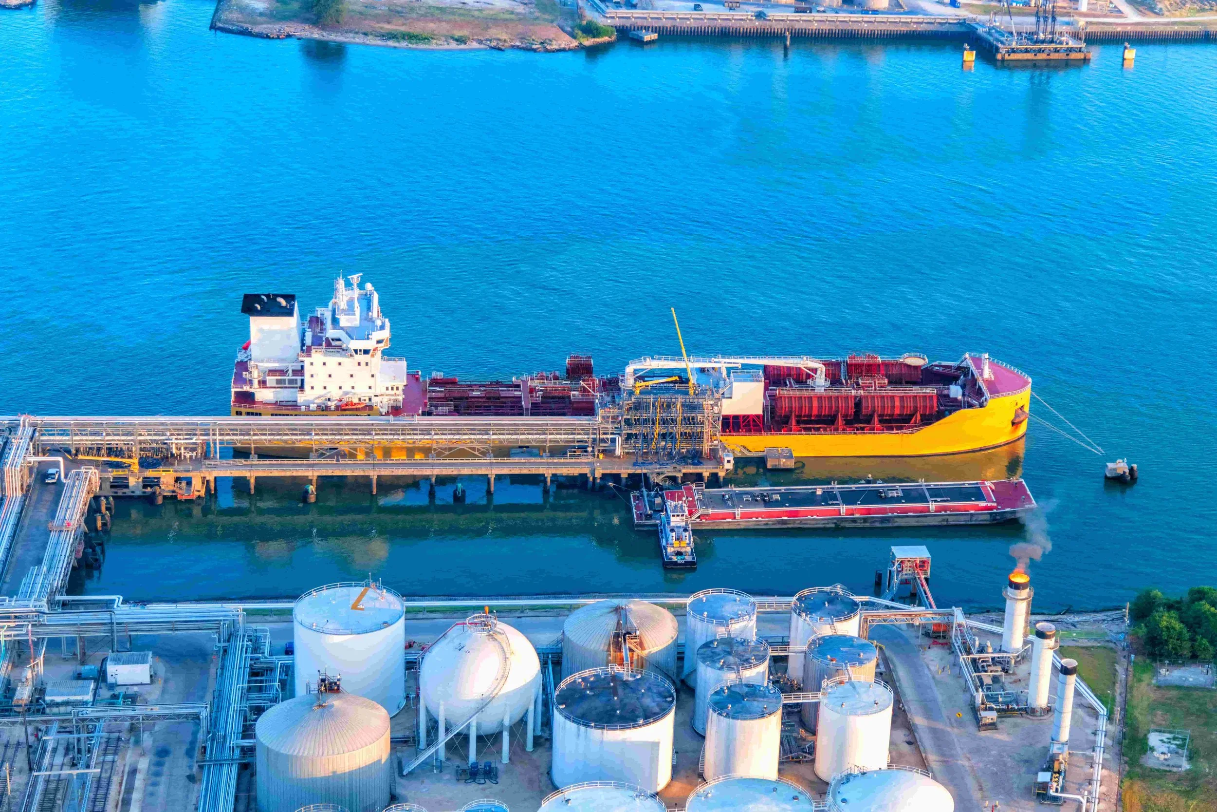 An aerial view of an industrial area with large storage tanks and a cargo ship docked at a pier in a harbor with clear blue water.