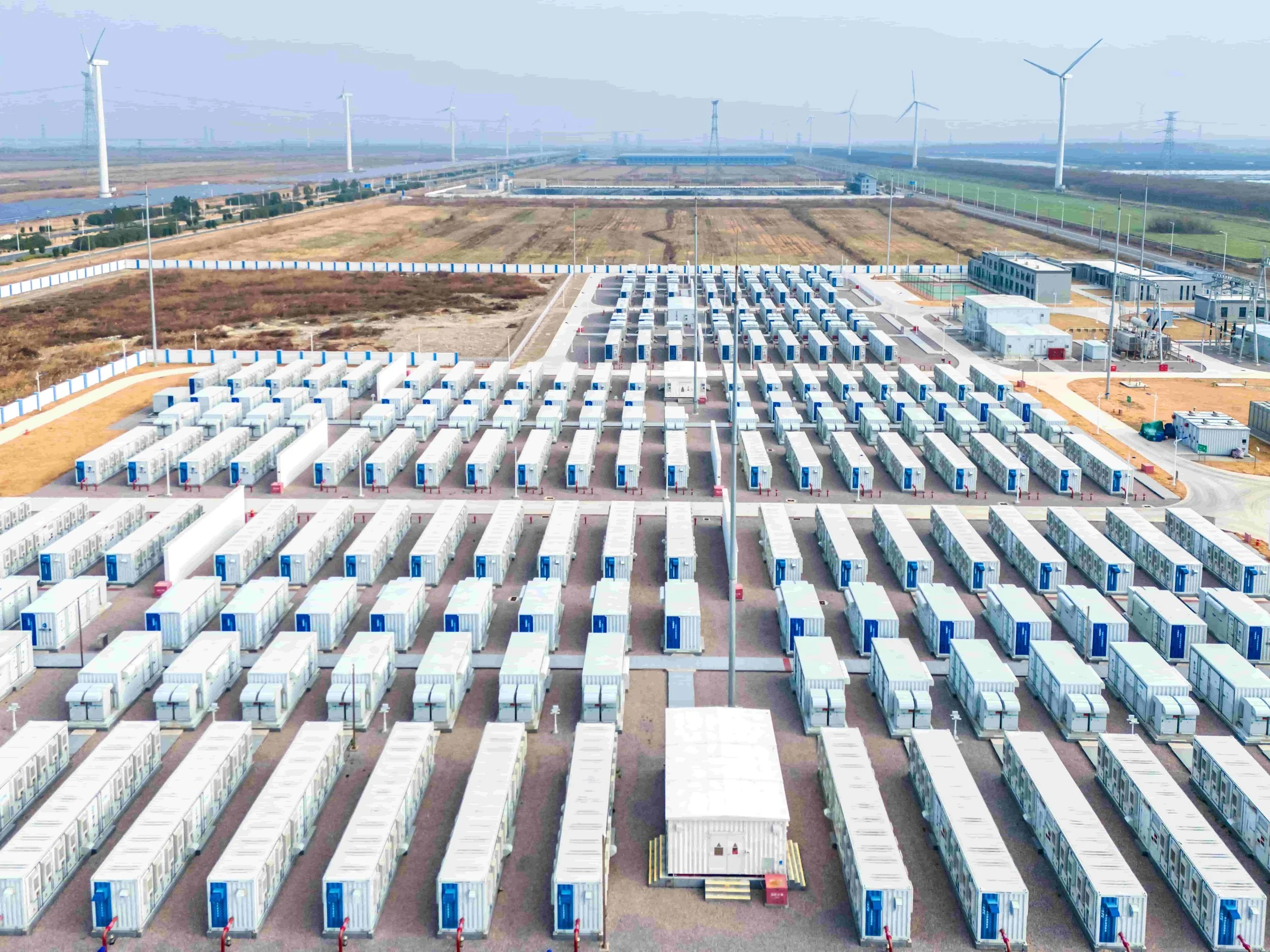 A large facility with rows of mobile storage units, wind turbines in the background, and a fenced perimeter, likely a data center or energy storage site.