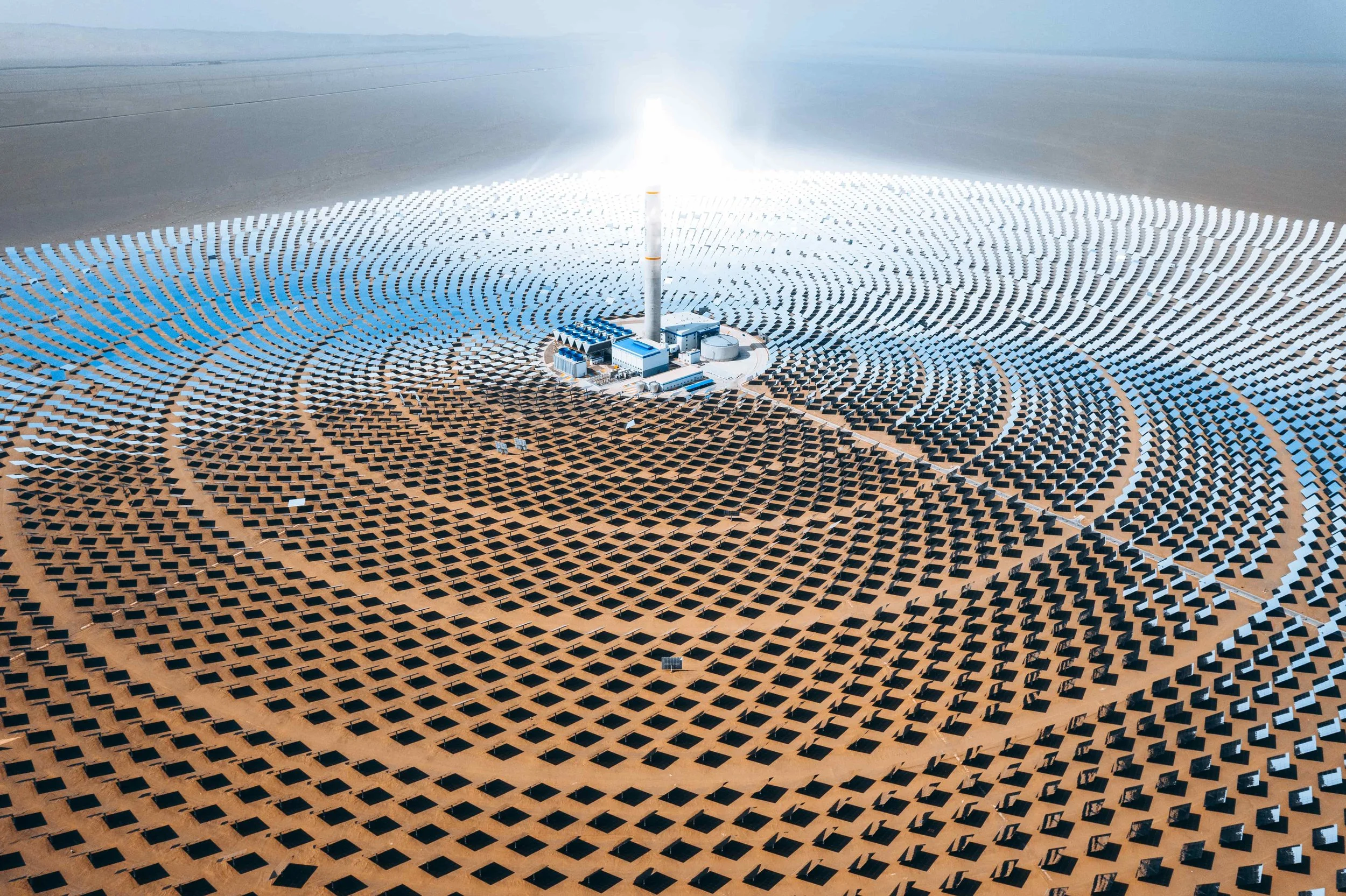 A solar power plant with numerous solar panels arranged in concentric circles around a central building with a tall chimney.