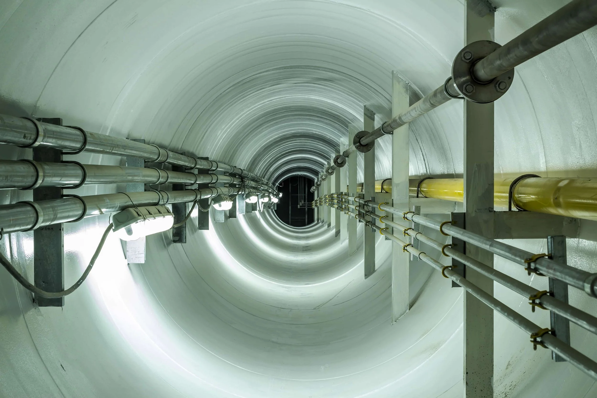 Looking down into a large, cylindrical, industrial pipe or tunnel with metallic pipes running along the sides and bright lights illuminating the interior.