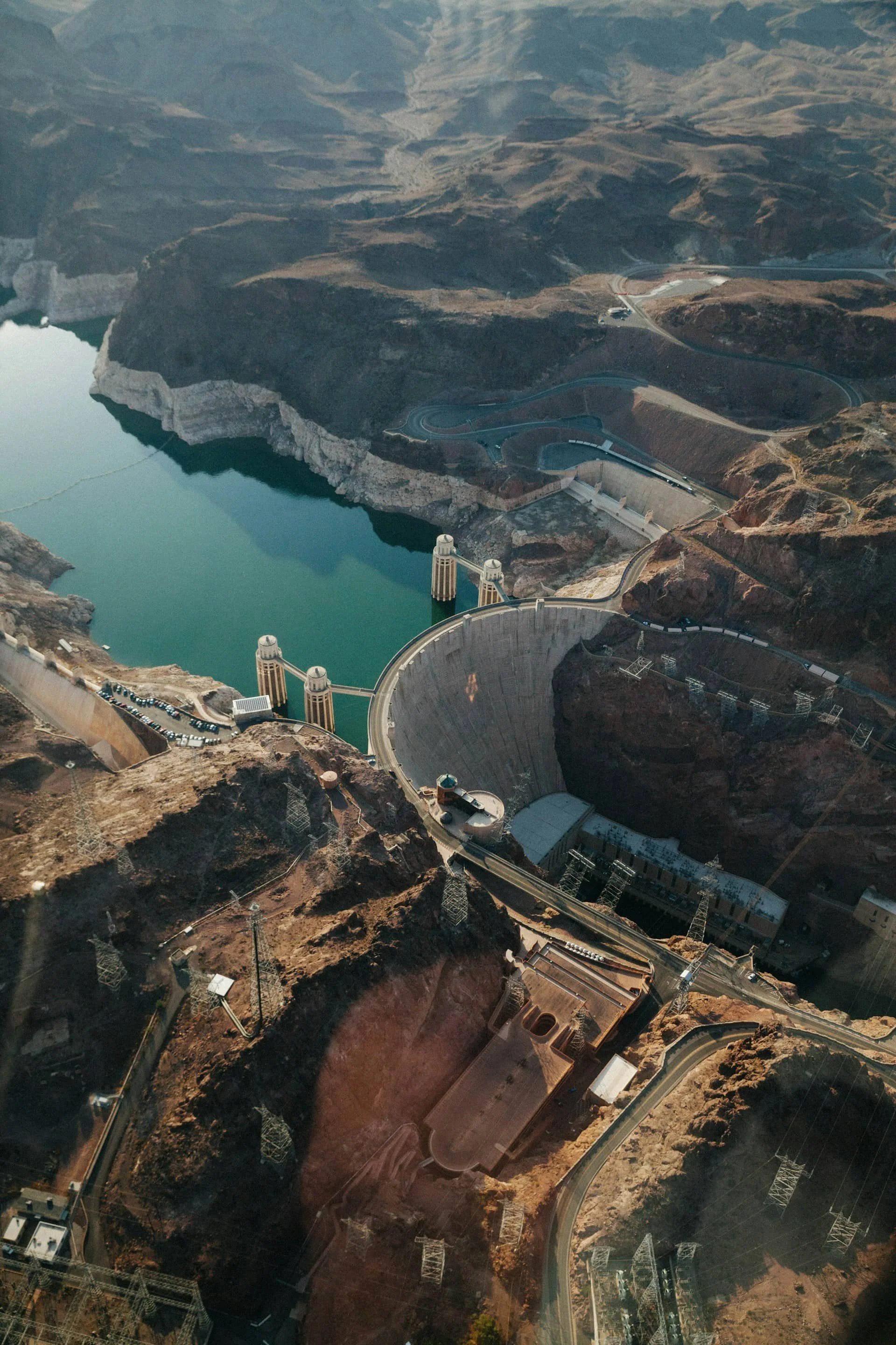 Aerial view of a large dam with a curved concrete structure, holding back water in a reservoir, surrounded by mountainous terrain and roads.