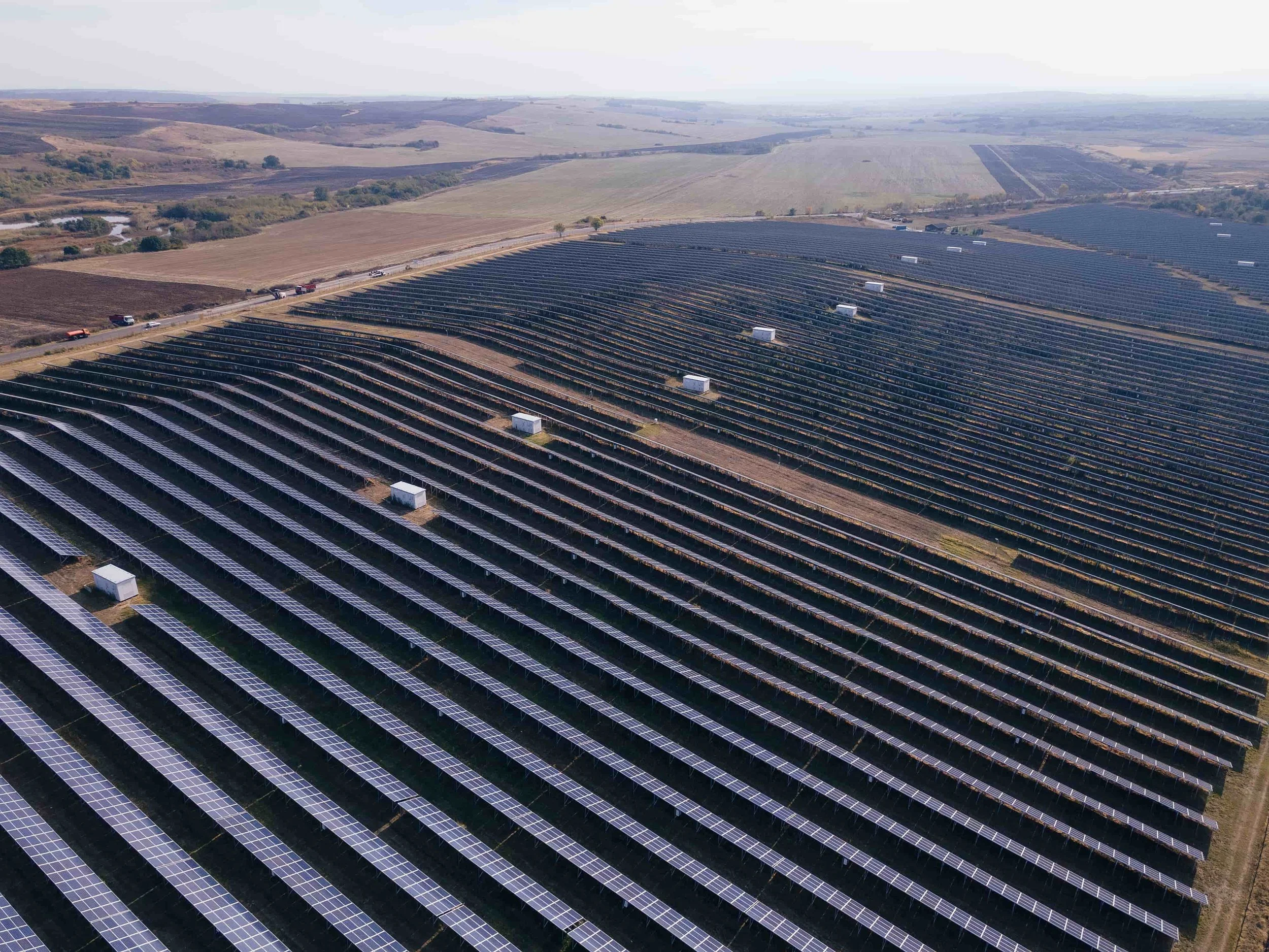 Aerial view of a large solar farm with rows of solar panels in a rural landscape with fields, hills, and a small river.