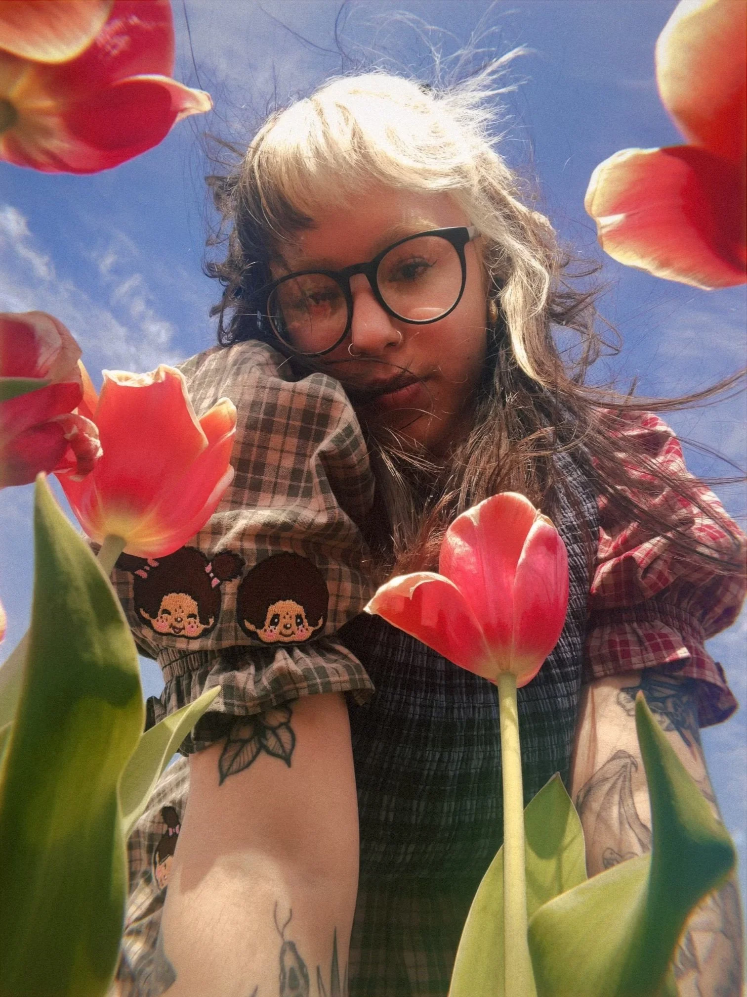 A young woman with blonde and black curly hair, wearing glasses and a nose ring, taking a selfie among pink tulips outdoors under a blue sky.