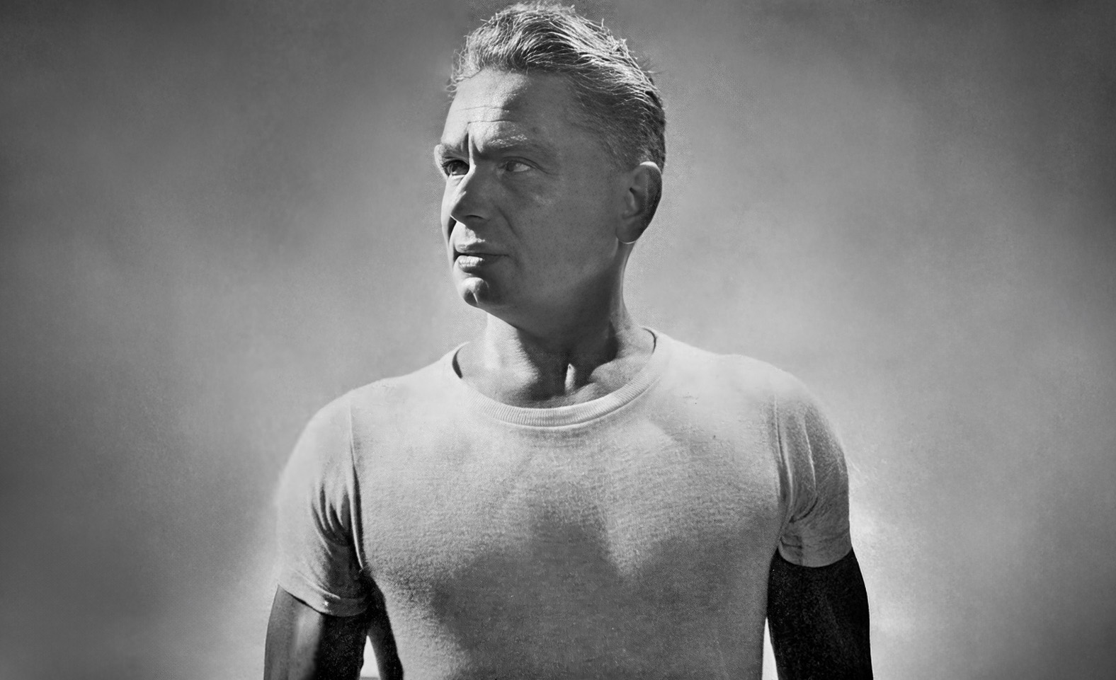 Black and white photograph of a man with short hair, wearing a plain t-shirt, standing against a plain background, looking to his left.
