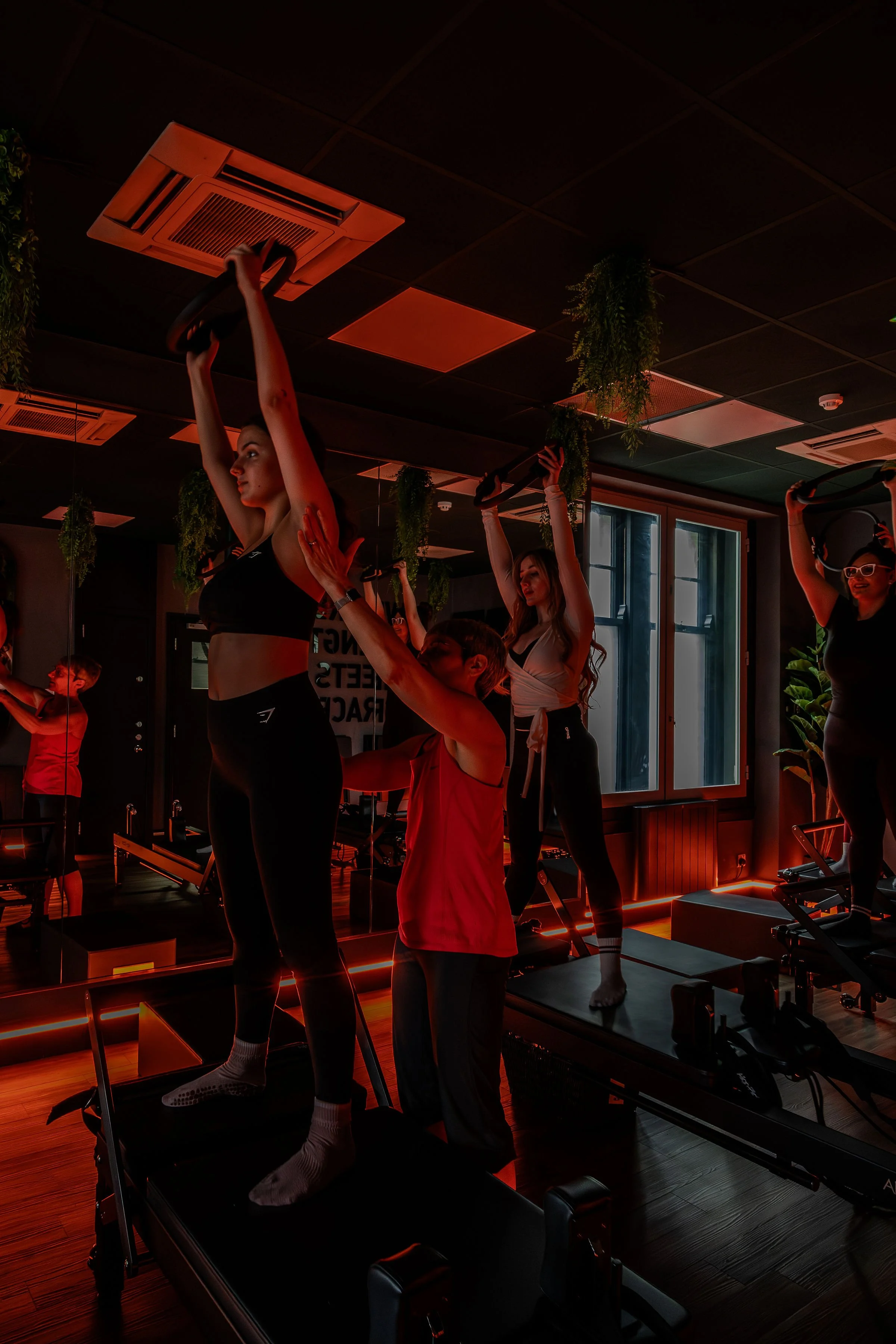 Women exercising on Pilates reformers in a fitness studio with black walls, mirrors, and hanging plants, illuminated by red lighting.