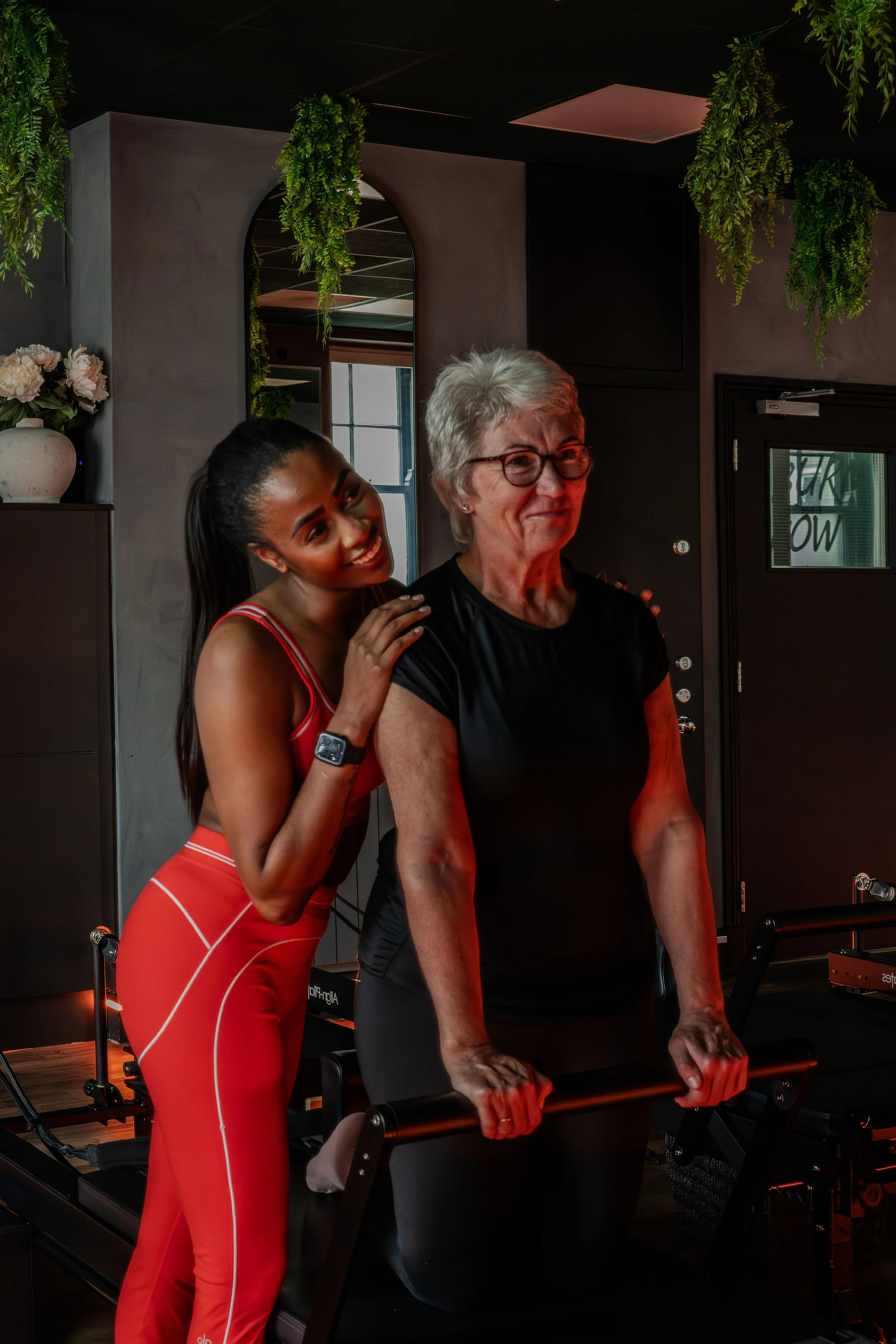 A younger woman with dark hair in a ponytail, wearing an orange workout outfit with white piping, hugging an older woman with short gray hair and glasses, who is dressed in a black shirt, in a fitness studio with dark walls and hanging greenery.