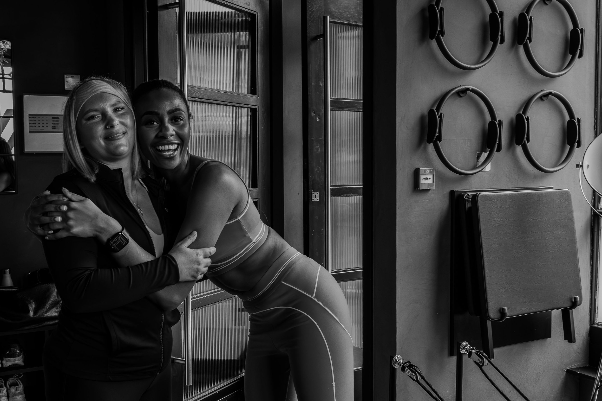 Two women hugging and smiling in a fitness or gym setting with exercise rings and fitness equipment on the wall.