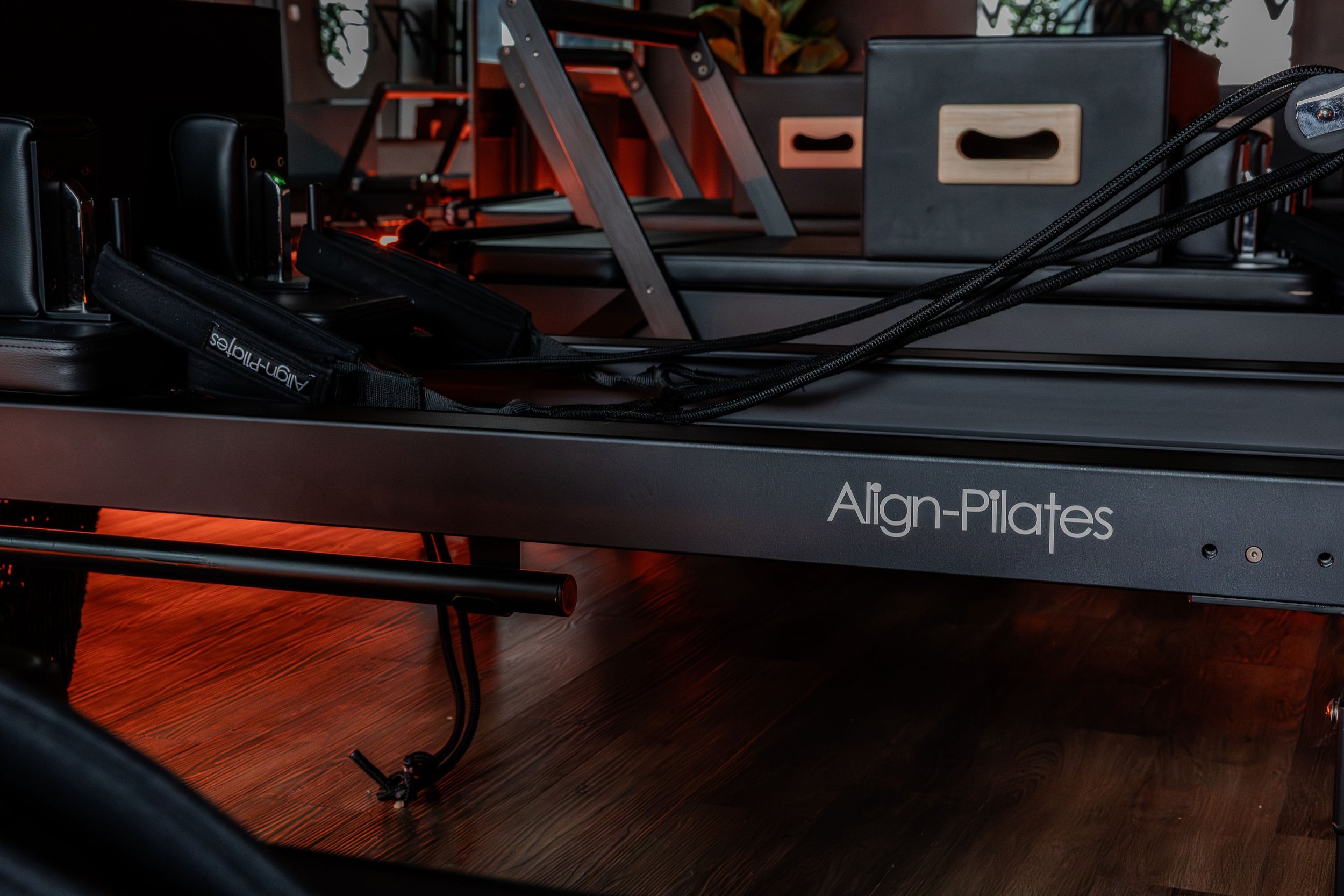 Exercise equipment on a wooden floor, including a black reformer with the words 'Align-Pilates' visible, and other fitness accessories.