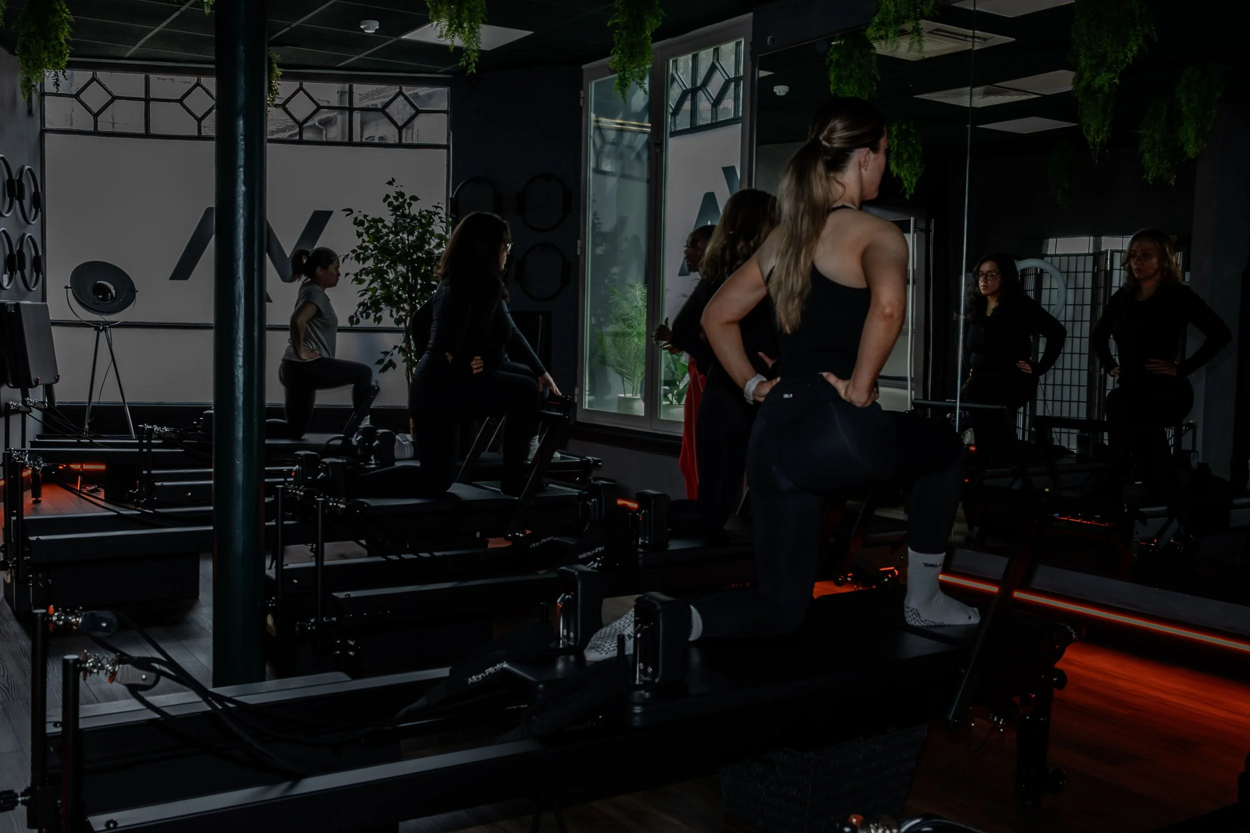 Group of women in a fitness class at a gym, with some standing and others sitting on exercise machines, in a modern gym with large windows and greenery outside.