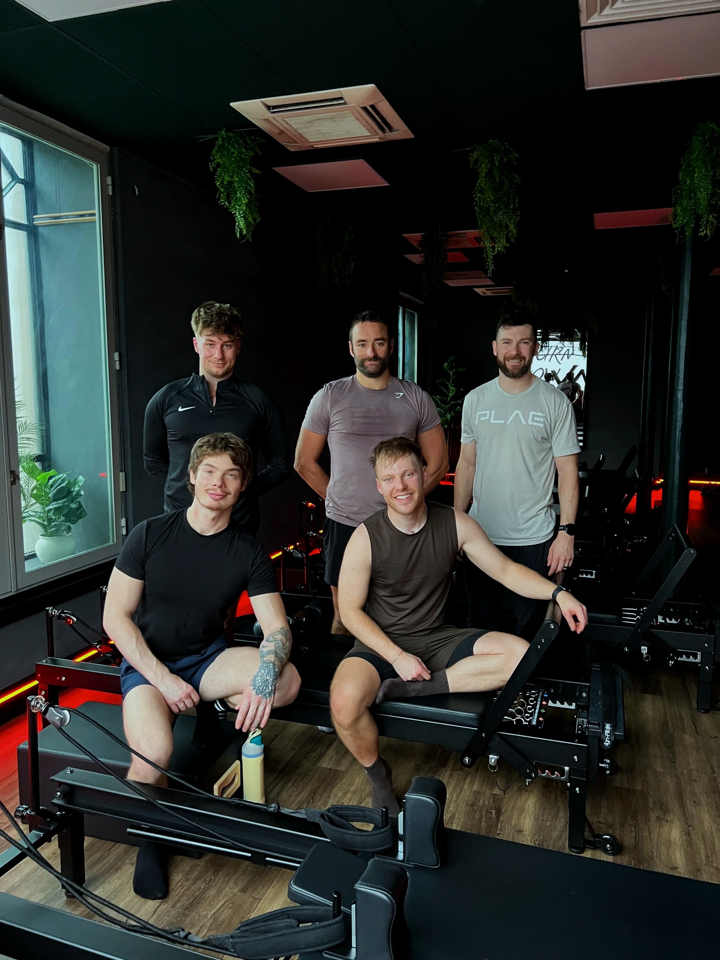 A group of five men in athletic clothes are gathered in a dark indoor space with fitness equipment and potted plants.