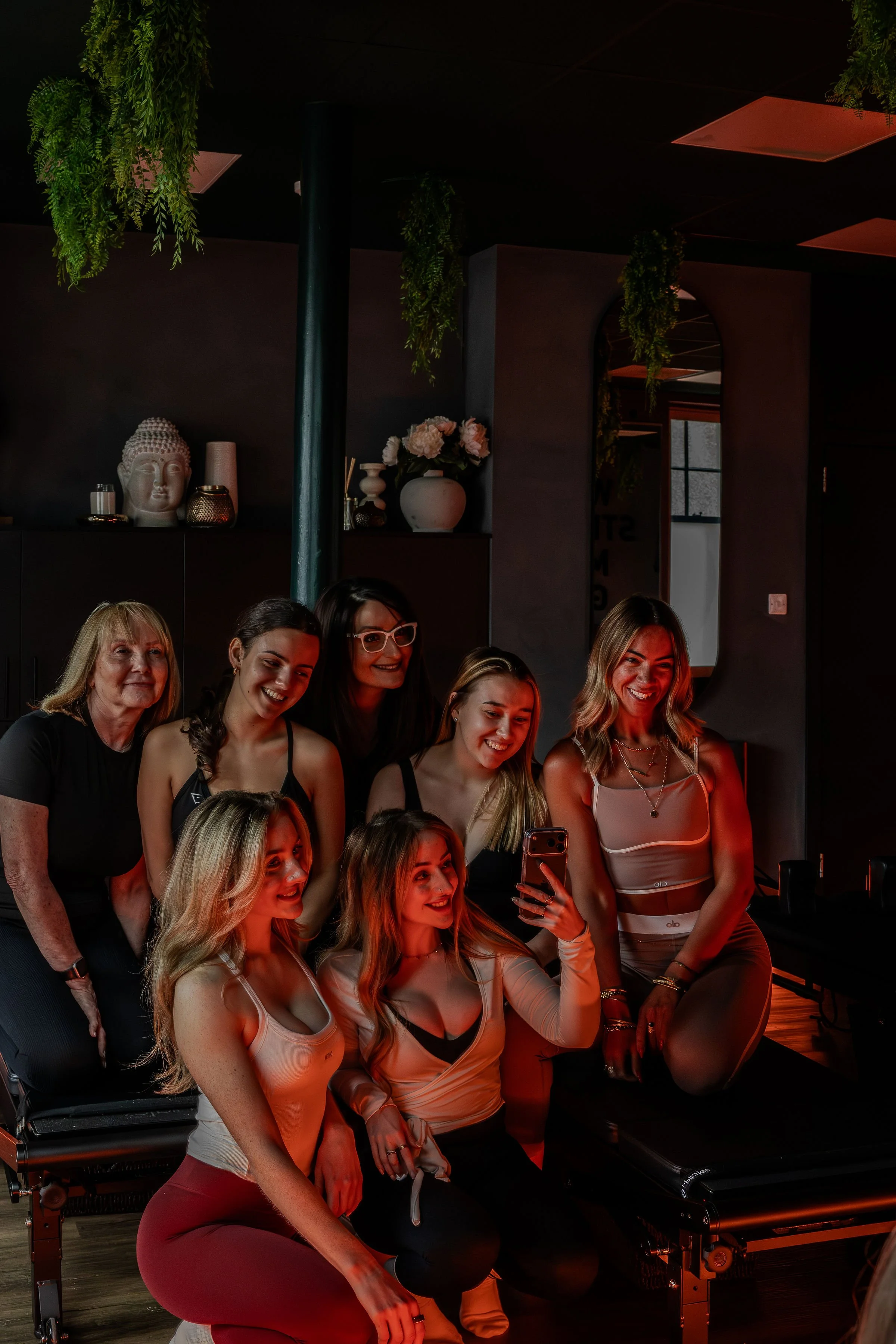 A group of women taking a selfie in a dimly lit room with dark walls decorated with hanging green plants, a Buddha head sculpture, and vases with flowers, with some women smiling and others looking at a phone.
