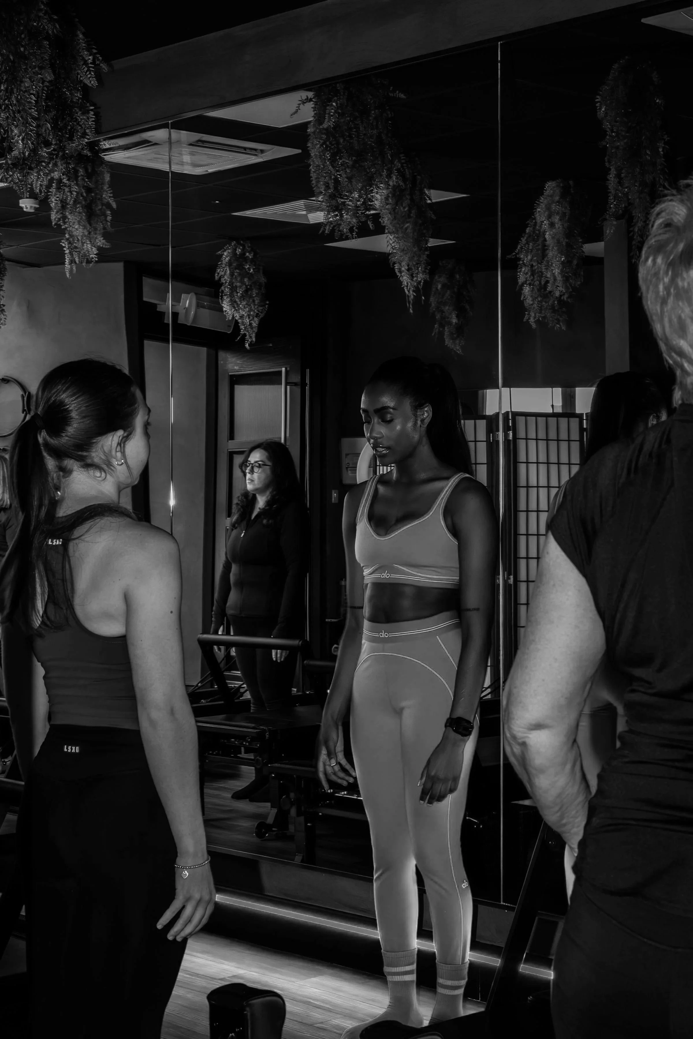 A group of women and men in workout clothes standing in front of a mirror, preparing for exercise in a gym with hanging plants and gym equipment visible.
