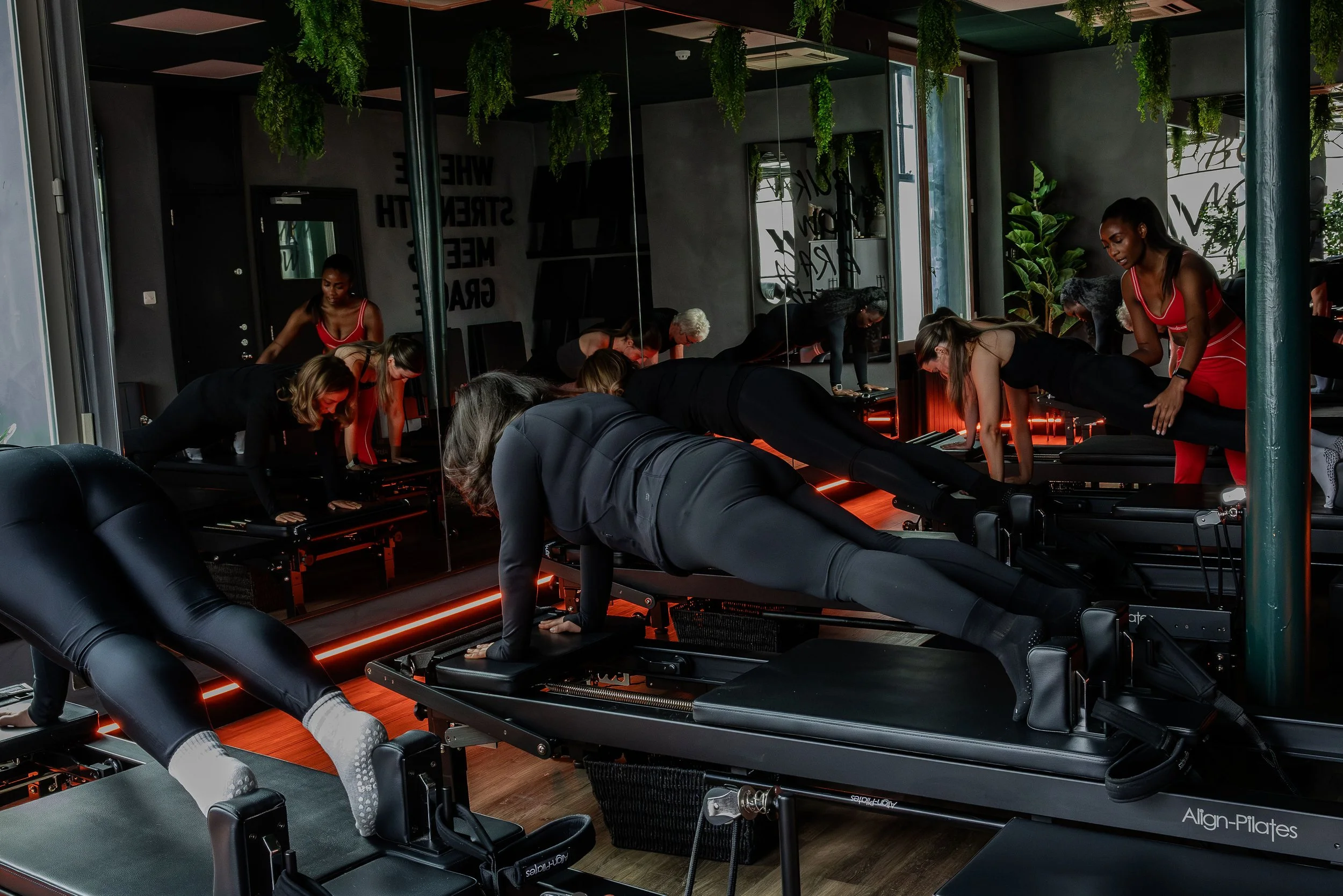 Group of people in a fitness class performing Pilates exercises on reformer machines in a modern, well-lit studio with plants and mirrors.