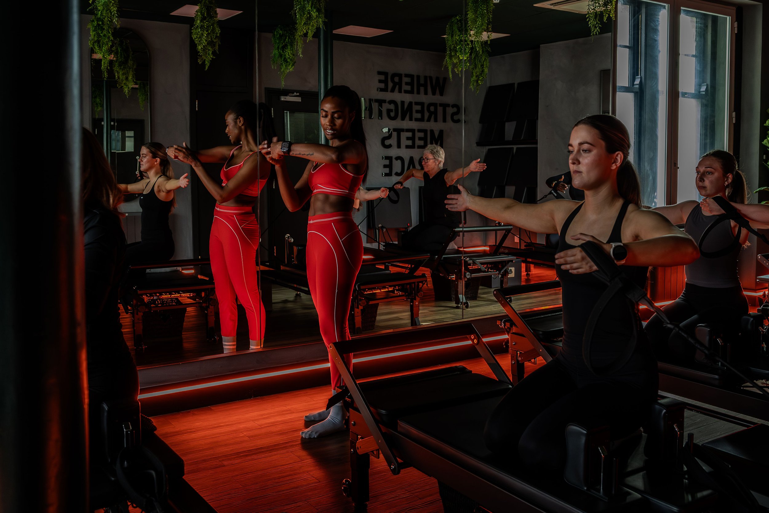 Group of women exercising on reformer machines in fitness studio with mirrors and hanging plants.
