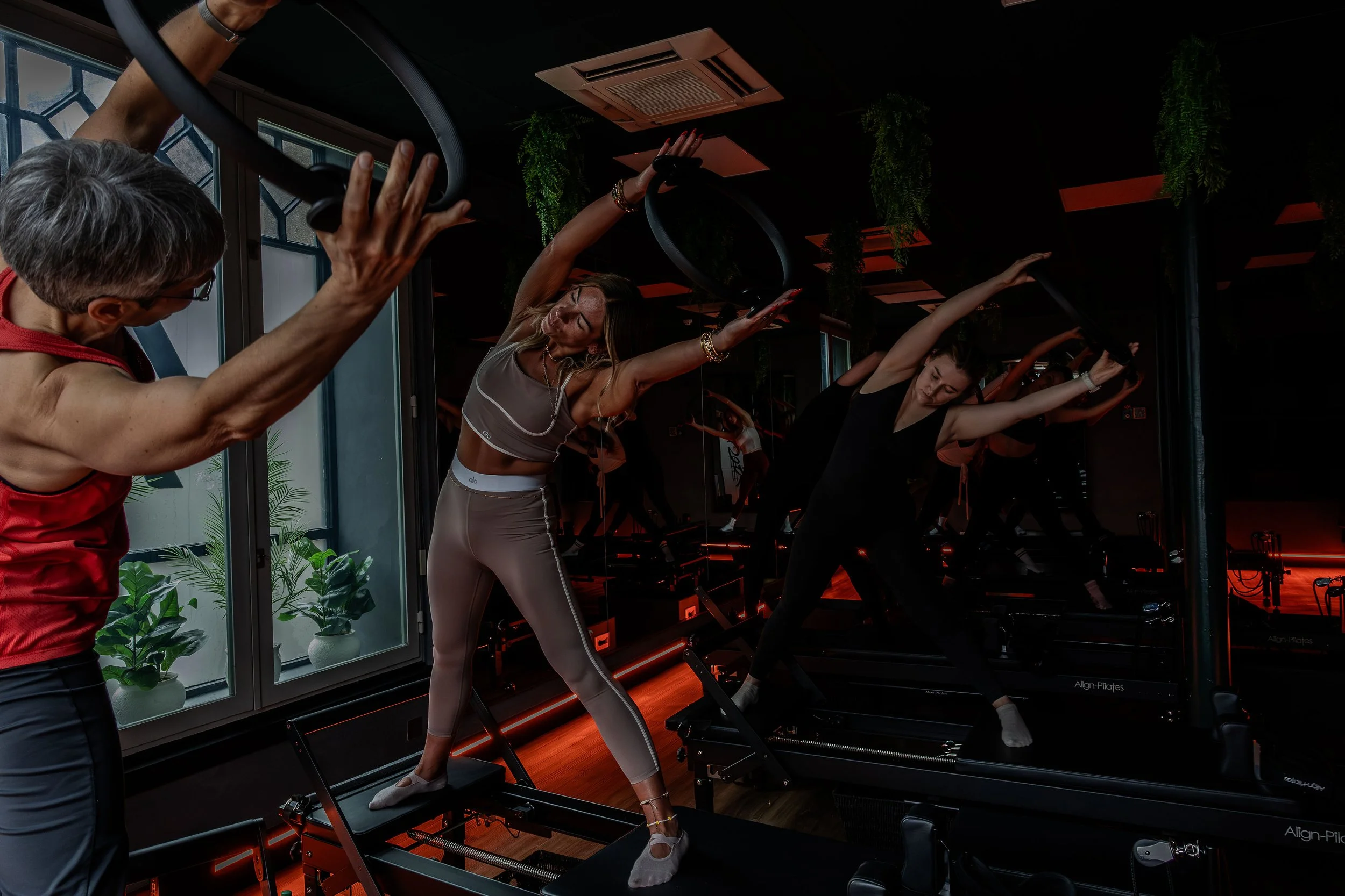 Women and instructor participating in a Pilates class using reformer machines in a studio with black walls and hanging plants