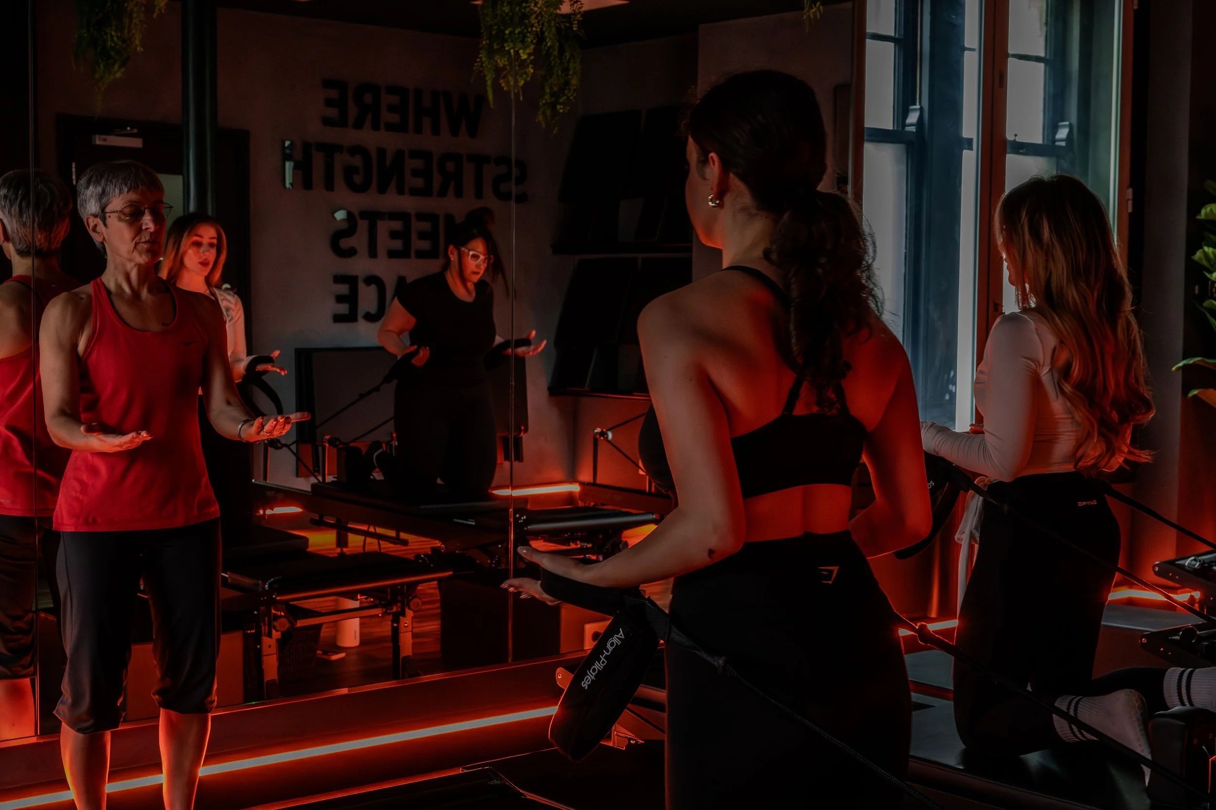 Group of women in workout attire in a fitness studio with mirrors and large windows, with warm lighting and plants.