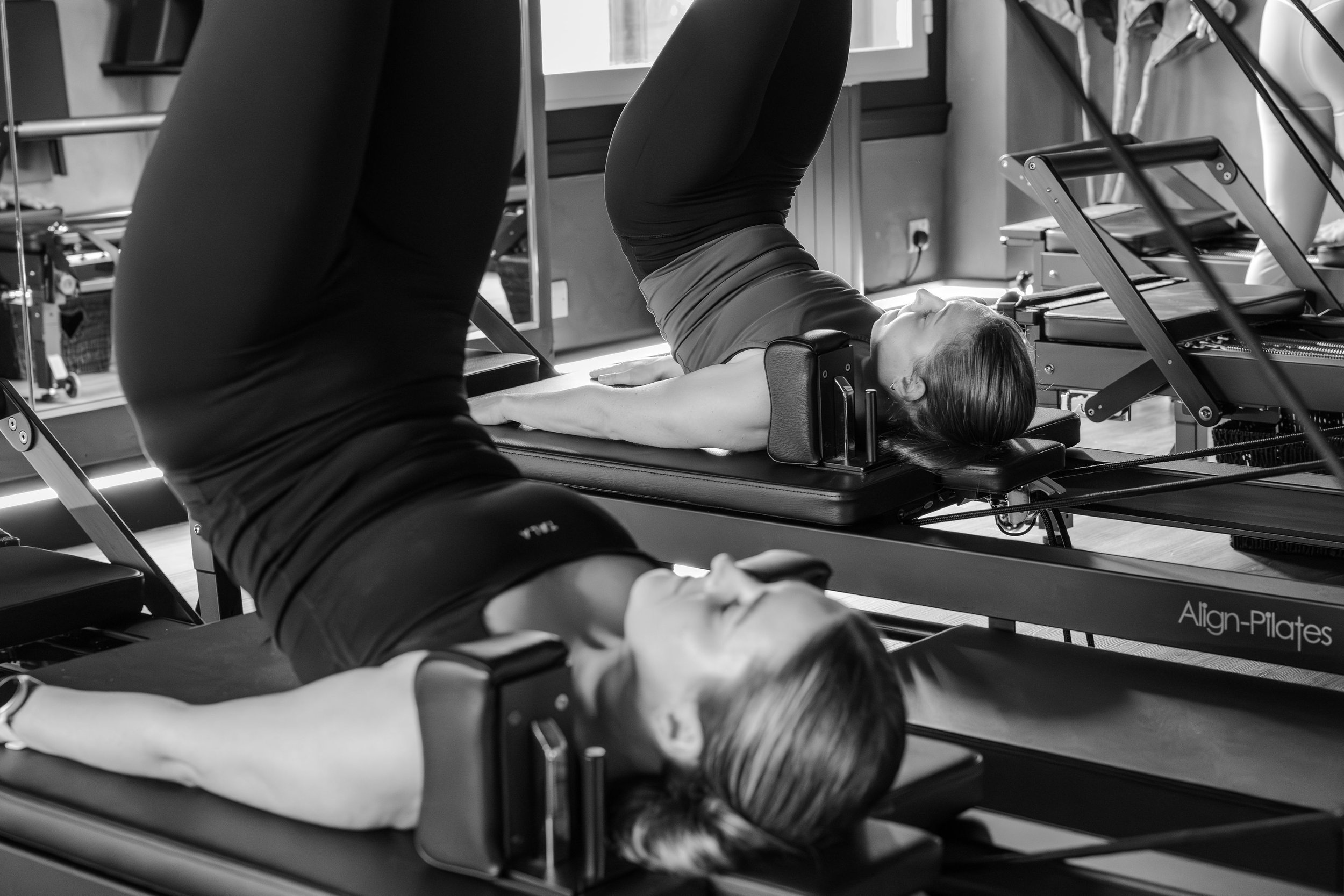 A woman lying on a Pilates reformer machine with her eyes closed, wearing workout attire, in a Pilates studio with equipment and windows in the background.