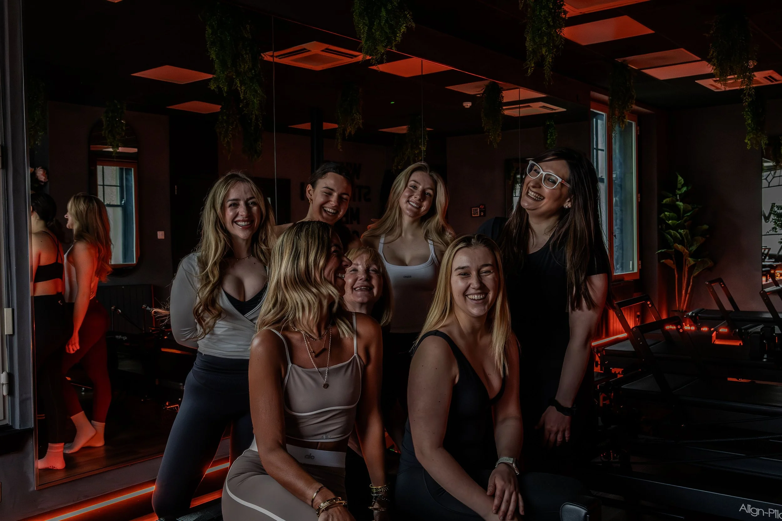 Group of women smiling and posing inside a fitness or dance studio with mirrors, plants, and fitness equipment.