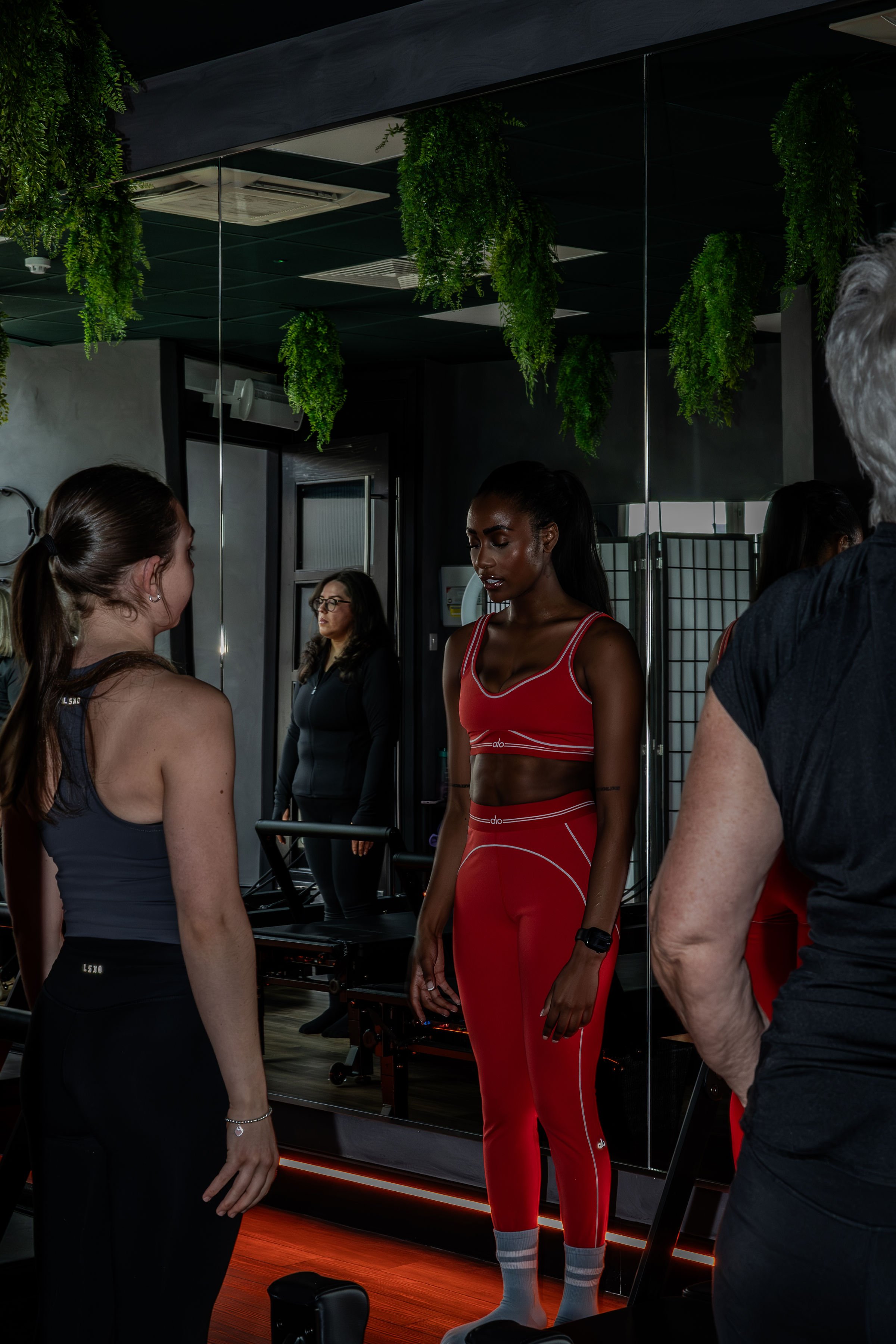 A woman in a red workout outfit standing in front of a mirror, surrounded by several people in a workout studio with green hanging plants on the ceiling.