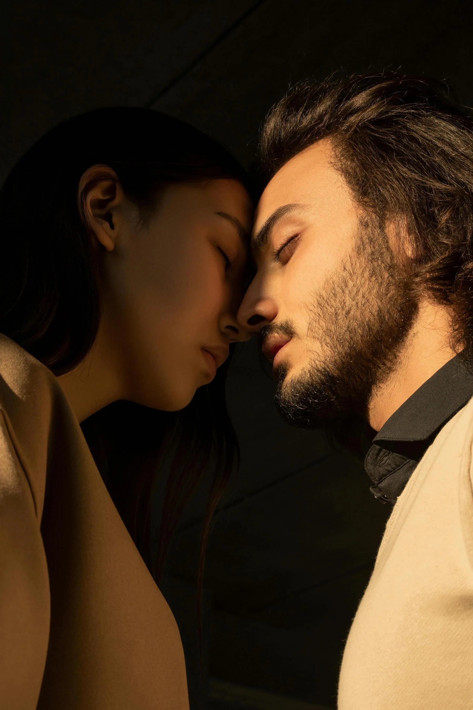 A close-up photo of a young woman and a young man with their foreheads touching, eyes closed, with warm lighting and dark background.