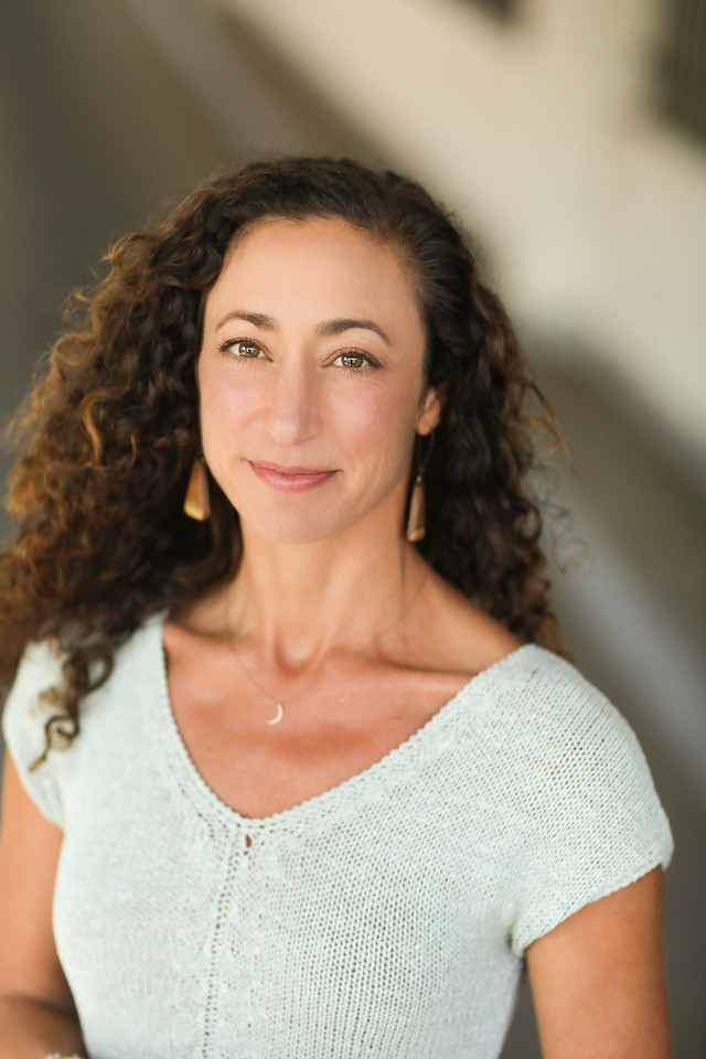 A woman with curly brown hair, light skin, and wearing a white knit top. She has a necklace with a small moon pendant and long earrings. She is smiling and looking at the camera.
