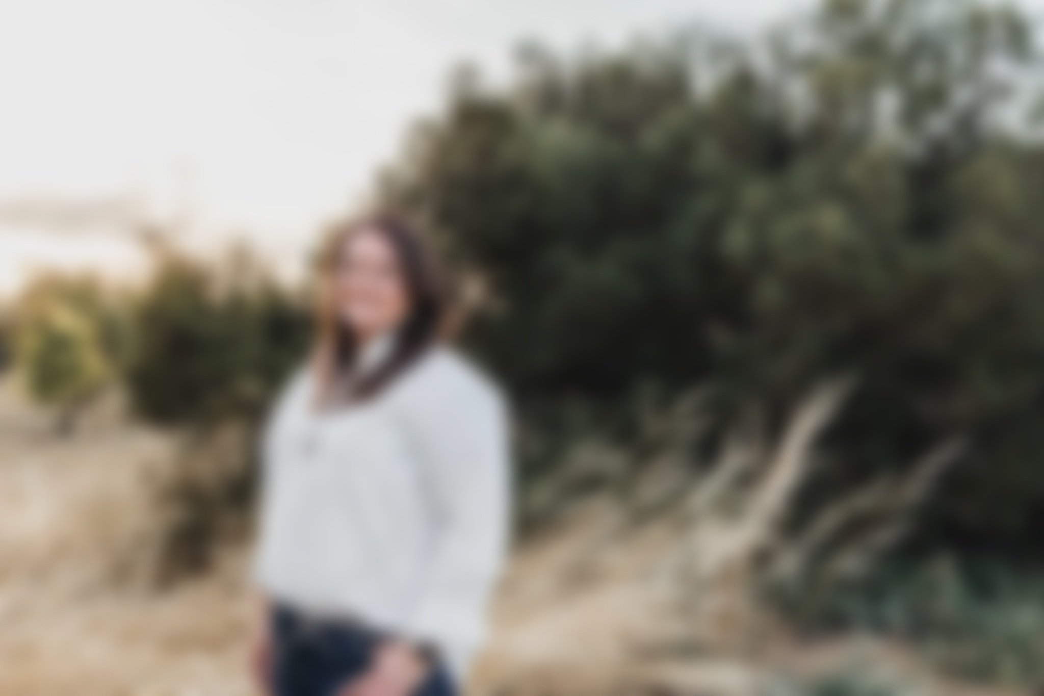 Blurred image of a woman with dark hair in a white dress standing outdoors near a large tree, with a clear sky and distant landscape background.