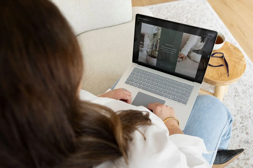 A woman sitting on a beige sofa using a laptop, with a wooden coffee table beside her holding a cup and glasses, in a cozy living room.