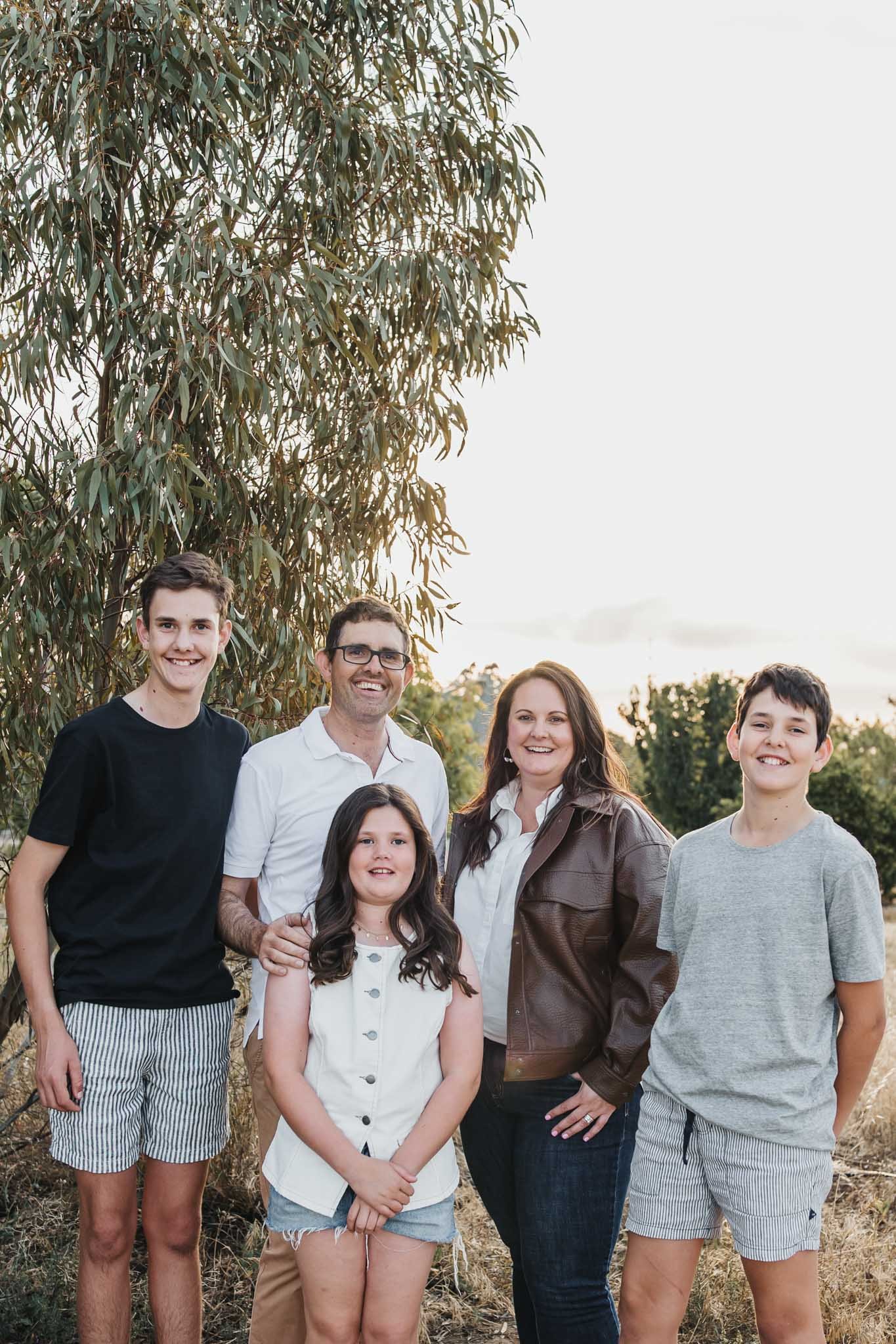 Group of eight people, including children and adults, standing outdoors near a tree, smiling at the camera during sunset.