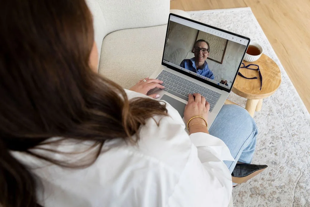 Woman in white shirt and jeans on a video call with a woman wearing glasses, sitting at a wooden table with a cup of tea and a pair of glasses.