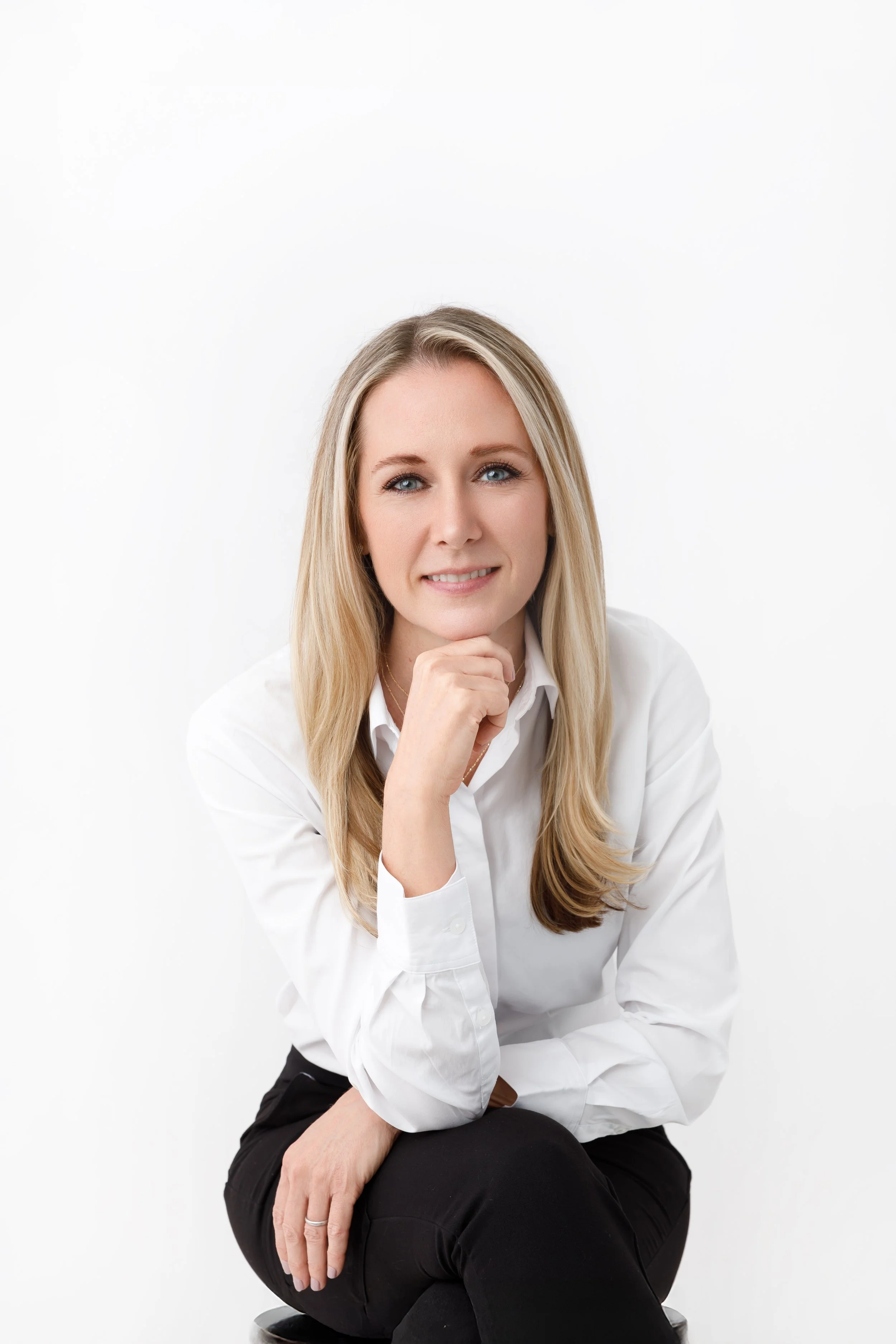 A woman with long blonde hair, blue eyes, and light skin, wearing a white button-up shirt and black pants, sitting in a relaxed pose with her chin resting on her hand, Skyiley Aesthetics Wellness in Sarasota, FL