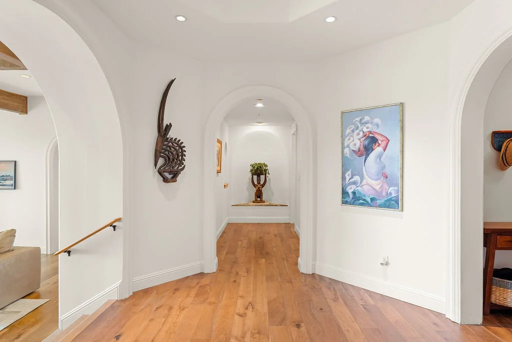 Hallway with white walls, hardwood floors, and arched doorways. Decorative art and a sculpture are displayed along the walls, with a potted plant at the end of the corridor.