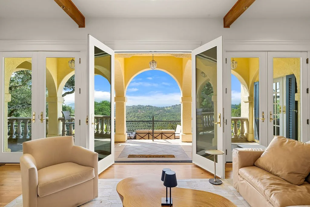 Inside a living room with open double glass doors leading to a balcony with yellow arches and a scenic view of trees and hills.