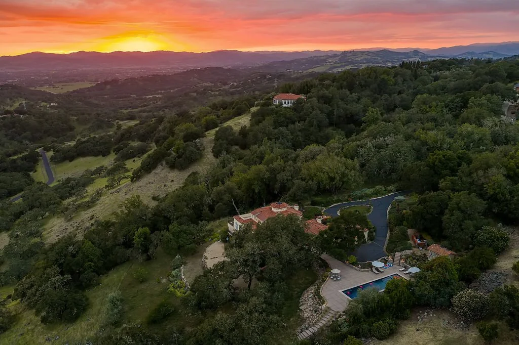 Aerial view of a hillside property with a swimming pool, surrounded by dense green trees and rolling hills during sunset.