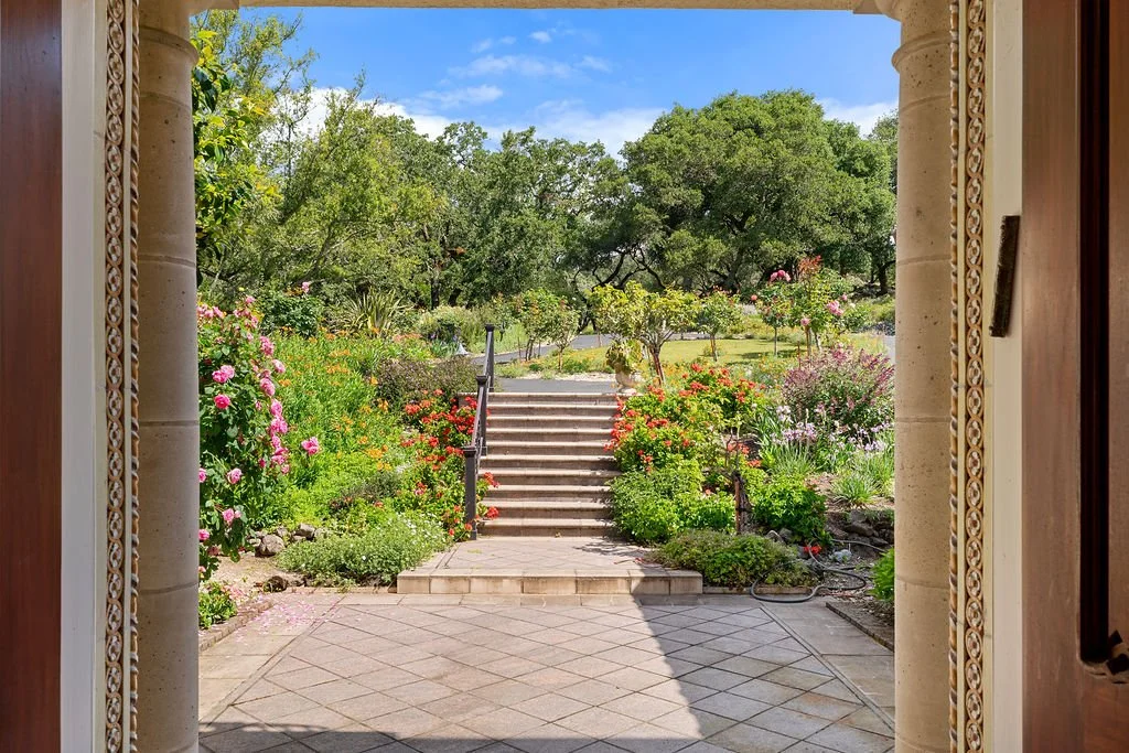 View of a vibrant, colorful garden seen from a doorway with stone columns, stone steps leading up to a lush green yard with various trees and flowering shrubs under a blue sky.