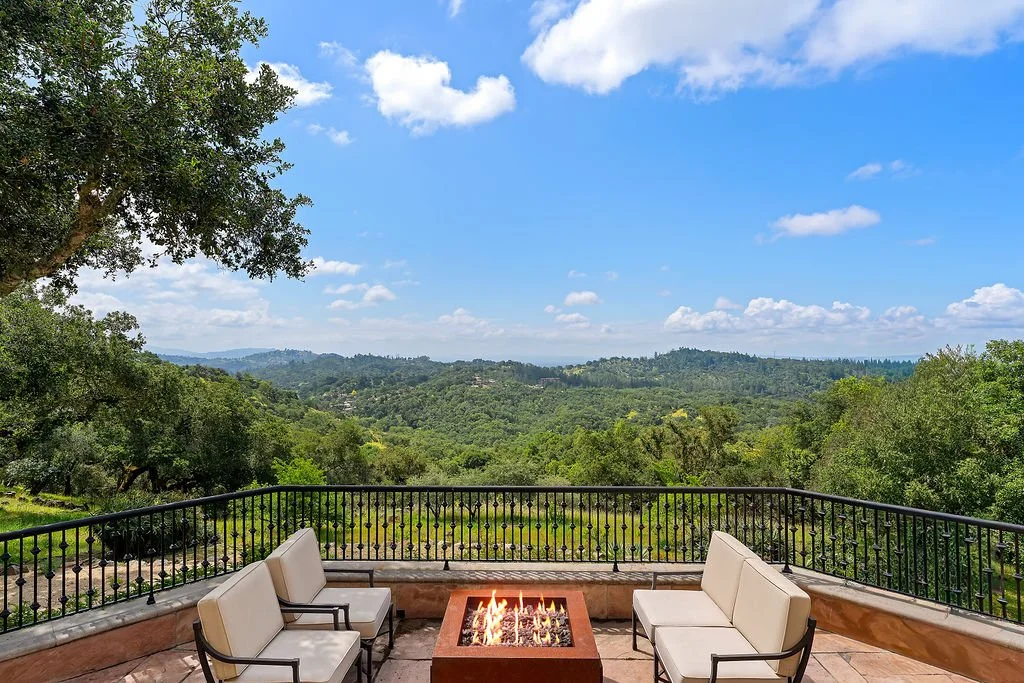 Balcony overlooking lush green hills and blue sky with clouds, with white outdoor chairs and a fire pit.