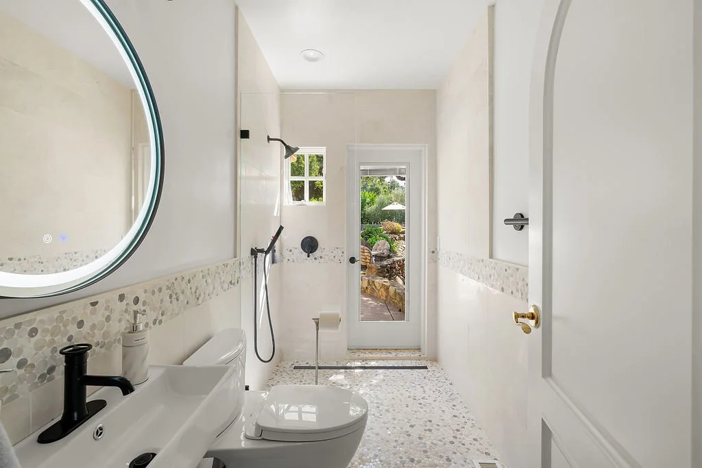 Modern bathroom with a round illuminated mirror, a white sink, a black faucet, and a toilet. The bathroom has light-colored walls with a pebble tile border, a shower area with a window, a handheld showerhead, and a glass door leading outside to a gar