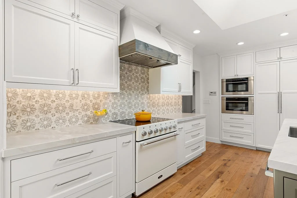 White kitchen with wood flooring, white cabinets, patterned backsplash, and built-in microwave and oven.