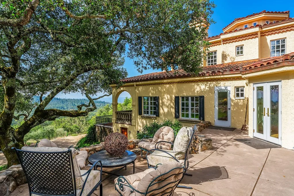 An outdoor patio with beige cushioned chairs and a table, shaded by a large tree, in front of a Mediterranean-style house with cream stucco walls, red-tiled roof, and blue shutters.
