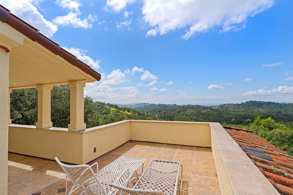 Terrace with two white lounge chairs overlooking a lush green landscape with rollers and hills under a blue sky with scattered clouds.