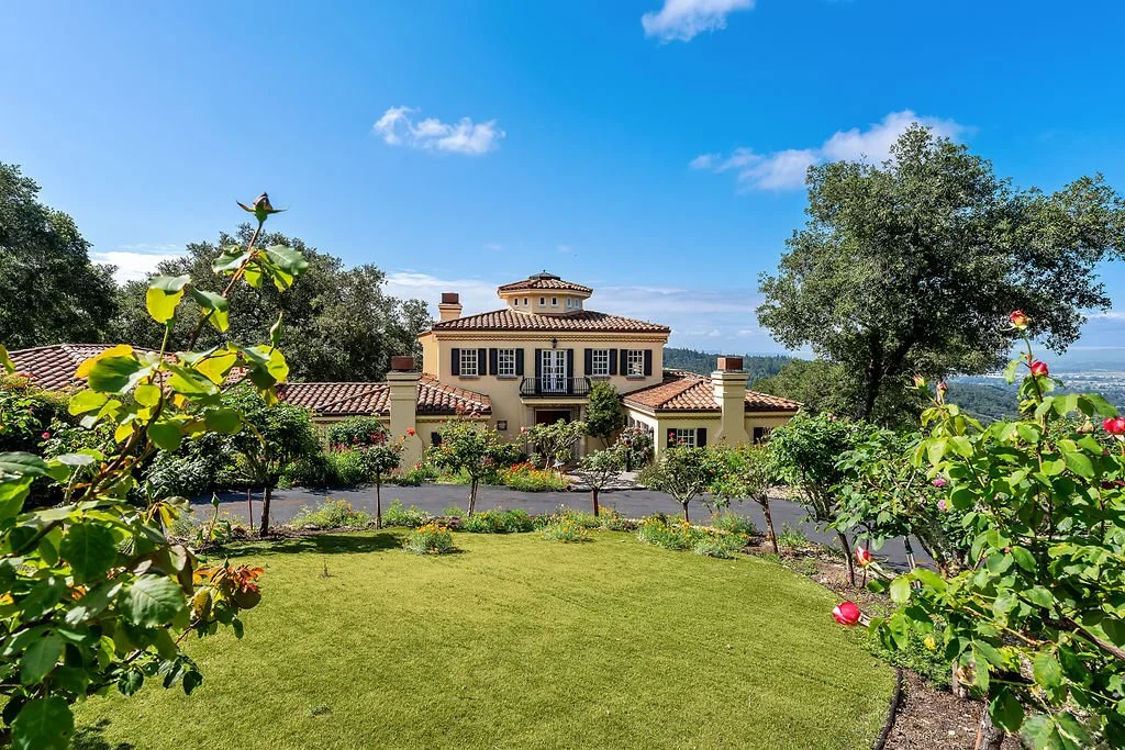 A large house with a red tiled roof surrounded by green trees and bushes, with a well-maintained lawn and a bright blue sky in the background.